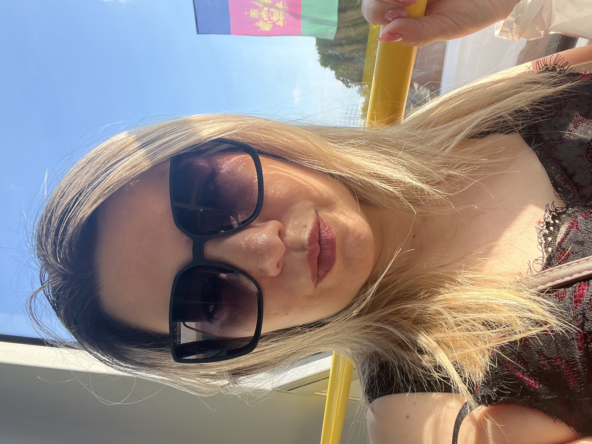 This is a selfie of a woman with blonde hair and sunglasses, taken outdoors on a sunny day. She is wearing a black and red lace top. In the background, a flag is visible, along with a yellow railing. The sky is blue and clear, suggesting it is daytime. The woman's expression is neutral. The image's orientation is rotated 90 degrees counter-clockwise.