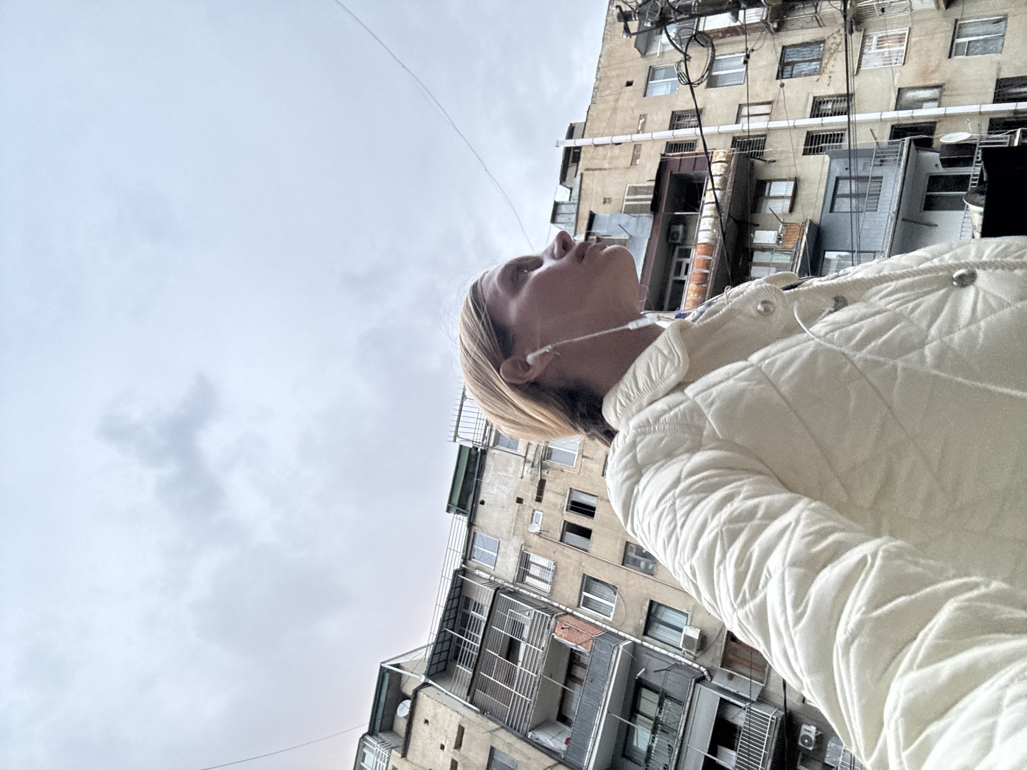 This low-angle photograph captures a woman looking upwards amidst a dense urban backdrop in Tbilisi, Georgia, under an overcast sky. The primary subject, a woman with blonde hair, is seen from the chest up in profile, her head tilted back as she gazes intently towards the sky or the upper reaches of the buildings. She wears a white quilted jacket and white wired earphones, suggesting she is listening to audio.
The setting features tall, multi-story buildings with an aged, concrete facade in a muted beige or dirty yellow hue. These structures are packed with numerous windows, some equipped with protective metal grates, and display various external features like air conditioning units, pipes, and a tangle of wires, indicative of a lived-in and somewhat cluttered urban environment. A thin, dark cable stretches diagonally across the cloudy sky, which dominates the upper-left and central portions of the frame, providing a sense of scale against the towering architecture.
The time of day is daytime, with bright yet diffused light due to the overcast weather conditions. The woman's expression is neutral and contemplative as she looks skyward. No specific event or strong emotion is depicted, but rather a moment of quiet observation within the bustling city. There is no visible text in the image.