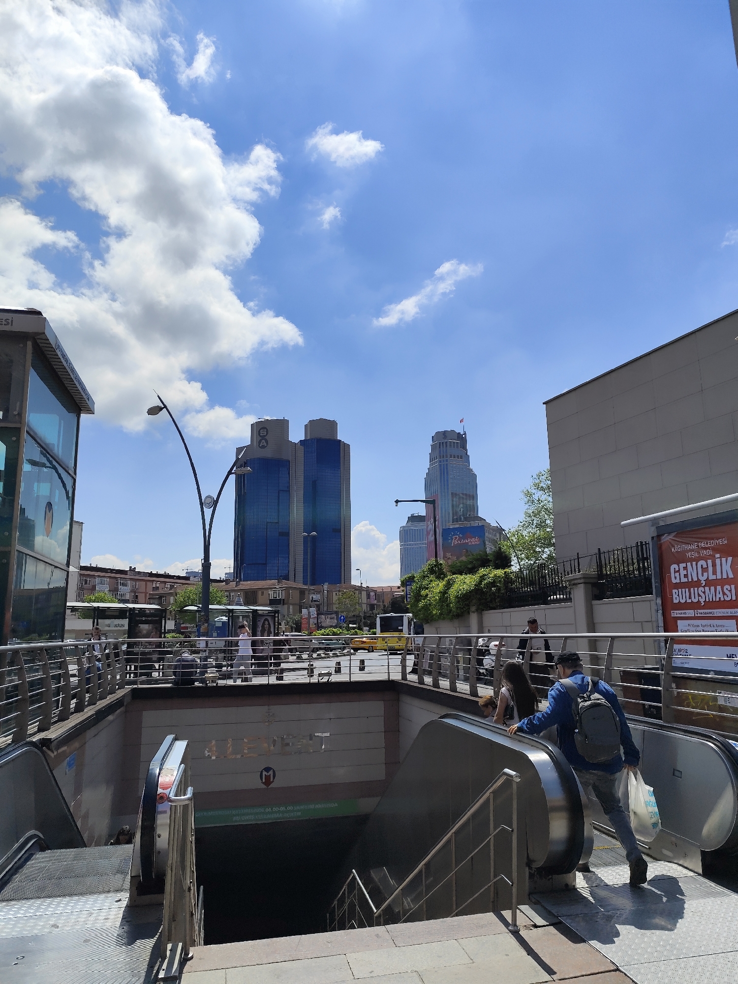 The image shows an urban scene in an unidentified city. The view is captured from an elevated position, likely the entrance of a metro station, where people are seen using an escalator to descend. The sky is a vibrant blue with scattered white clouds. In the background, there are modern buildings, including high-rise towers made of glass and steel. Streetlights with curved arms are also visible. Across the street, there are bus stops and signs of pedestrian activity. To the right of the frame is a wall with an advertisement poster.