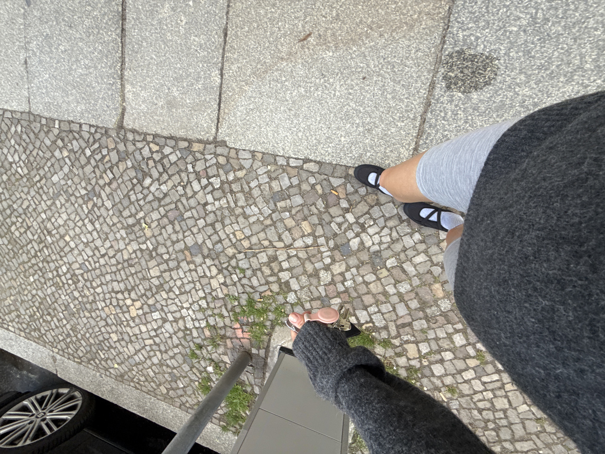 A first-person, high-angle shot depicts a person standing on a sidewalk in Berlin, Germany. The sidewalk is composed of two types of stone: a section of large, rectangular, light gray slabs, and an adjacent area of small, square cobblestones. The person is wearing gray shorts, white socks, and black Mary Jane shoes with white trim. They also wear a dark gray, oversized sweater.
The person is holding a set of keys with a light pink, circular fob attached. A gray metal pole is visible, along with a partially obscured gray and black object. A black car with silver rims is partially visible in the lower left corner. Patches of green grass grow between the cobblestones.