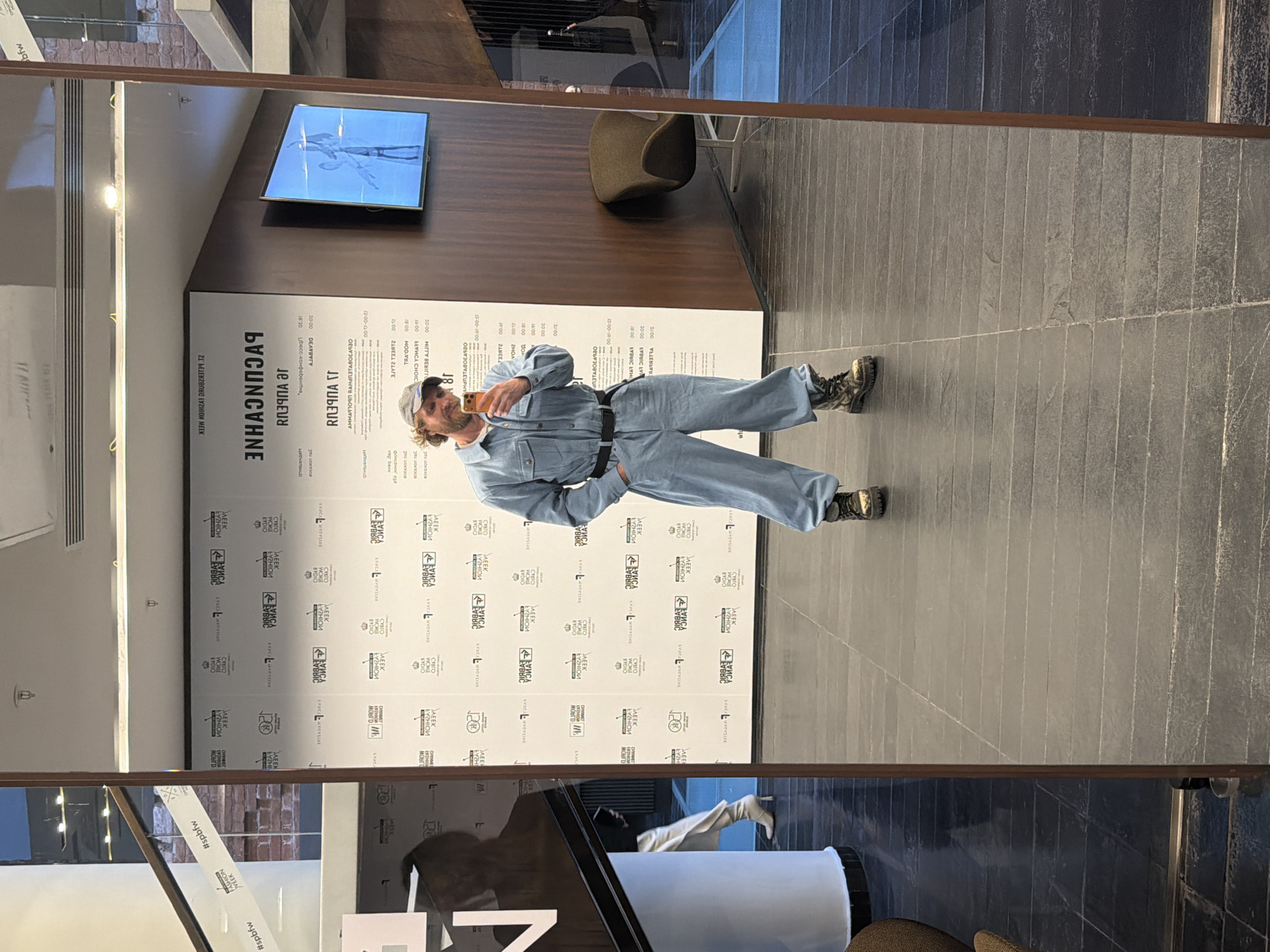 An indoor scene in what appears to be a public building or exhibition space, likely in Saint Petersburg, Russia, given the visible text and context. The primary focus is a full-body reflection of a person in a large, prominent mirror. This individual, a man with a beard and a cap, is wearing a light blue denim jumpsuit or overalls and sturdy, dark boots. He is holding a mobile phone, possibly taking a selfie or checking his appearance, and displays a slight smile or smirk.
Behind the subject, a large white information board is visible, featuring text primarily in Russian Cyrillic. Un-mirrored text includes "РАСПИСАНИЕ" (SCHEDULE) and "Лекции" (Lectures), alongside various times and names such as "ДЕВРИШ", "АНАТОЛИЙ", and "ЕЛЕНА ЧОЙСИ", indicating event details or speakers. Numerous logos incorporating the words "WEEK" and "FASHION" are printed on the board, and a hashtag "#spbfw" (Saint Petersburg Fashion Week) is visible in another reflection, strongly suggesting the setting is related to a fashion event. A television screen displaying an image of a person is mounted on a dark wooden panel wall section. The floor consists of long, narrow gray tiles. Partial reflections of other people and objects, including a contemporary upholstered chair, are visible within the mirror, indicating a lively, well-lit indoor environment.