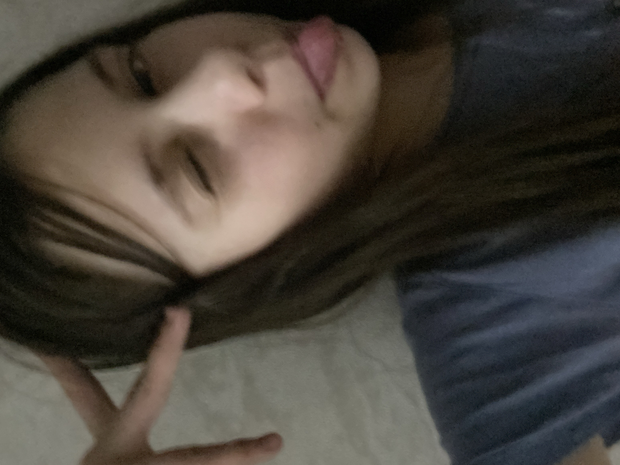 A person with brown hair is lying down, sticking their tongue out, and making a peace sign. The person is lying on what appears to be a bed with a light-colored sheet and is wearing a blue shirt. The location of the image is not discernible.