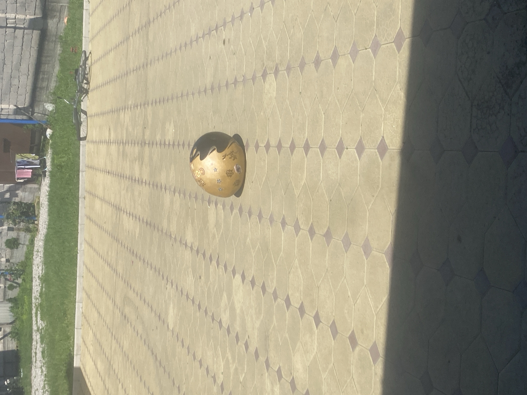 The image shows a paved courtyard with a patterned surface, likely in Belaya Rechka, Russia. A single, light-colored sphere with dark markings rests on the paving stones. To the right, a bicycle lies on its side. In the background, there is a patch of green grass, a building, and a clothesline with laundry. A shadow falls across the foreground of the image.