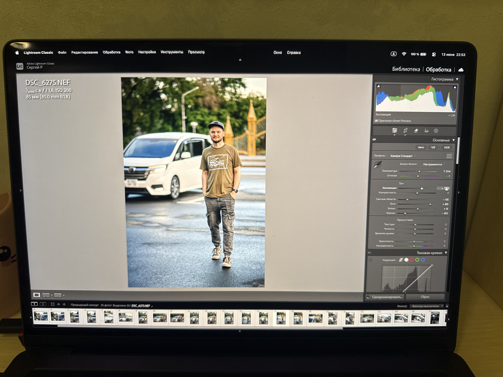 The image shows a laptop screen displaying Adobe Lightroom Classic software. The main subject is a photograph of a man walking on a wet street, with a white car and two golden spires in the background. The man is wearing a t-shirt, cargo pants, and a baseball cap. The Lightroom interface is visible on the right side of the screen, showing editing tools and sliders for adjusting image properties. A filmstrip of other photos is displayed at the bottom of the screen. The date and time displayed in the software are 13 июня 22:53. There is no visible indication of the image being taken in Kurgan, Russia.
