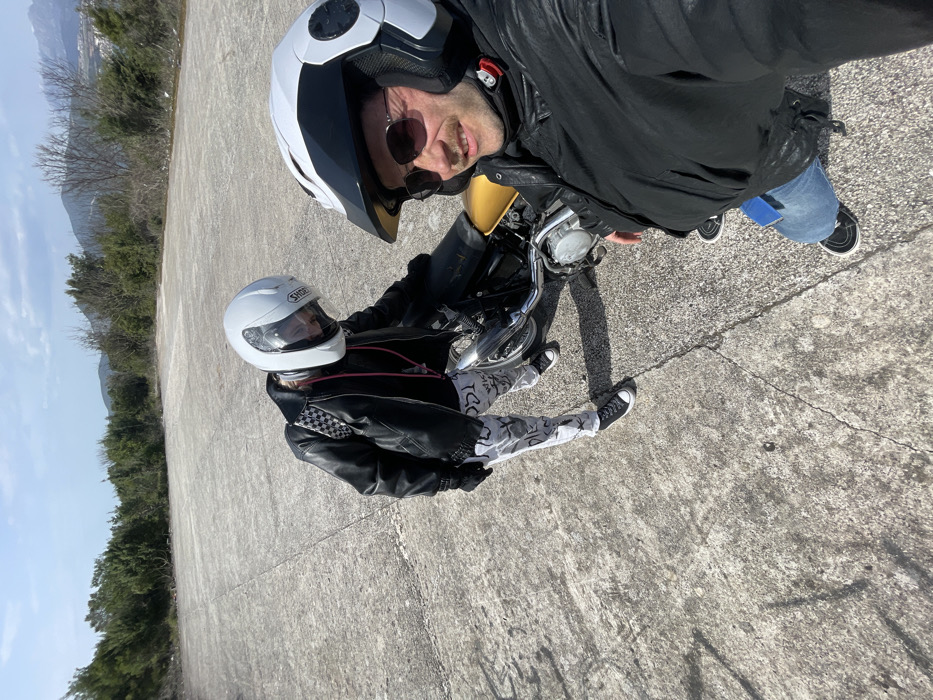 Two individuals in motorcycle gear are captured on a large, cracked concrete slab in Bigova, Montenegro, under a partly cloudy sky. In the foreground, a male with a mustache wears a white and black modular motorcycle helmet, dark sunglasses, a black leather jacket, blue jeans, and dark sneakers. He holds the camera, taking a selfie. Behind him, positioned to the left, a second person wears a white Shoei full-face helmet, a black leather jacket, black gloves, and white pants with dark graphic patterns. A black and white checkered backpack is on their back, and a pink lanyard is visible under their jacket. This individual stands next to a motorcycle featuring a yellow fuel tank and chrome components. The concrete slab is bordered by green Mediterranean vegetation, with distant hills and mountains visible under the blue sky. White buildings are discernible on a distant hillside.