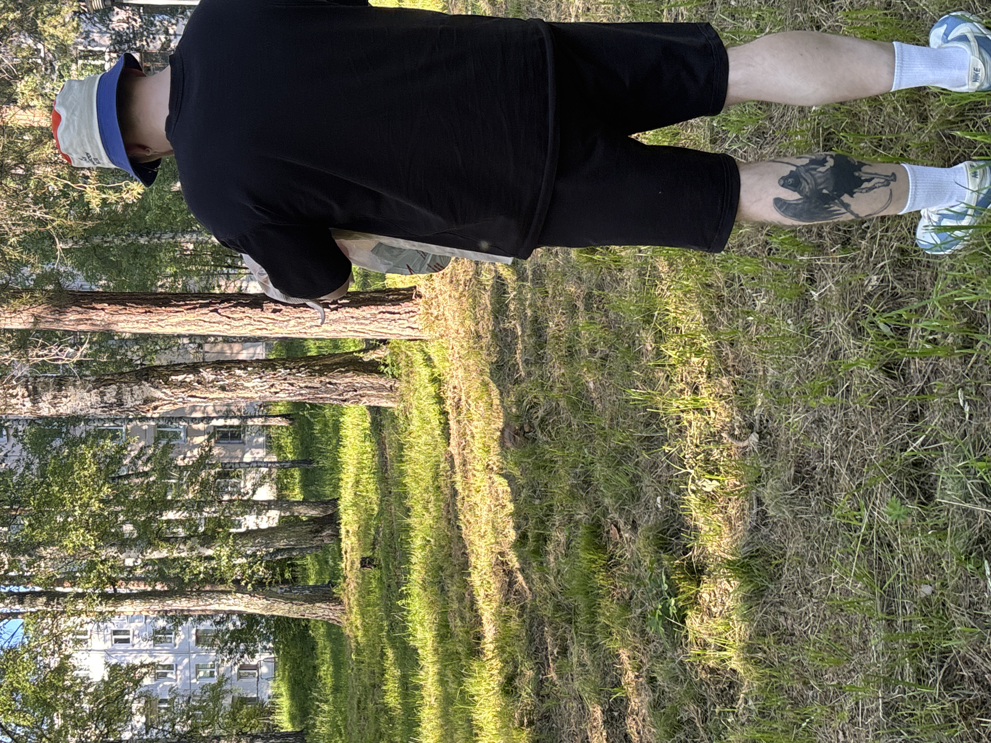 A person, seen from the back and slightly from the side, stands in a grassy area with trees. They are wearing a black t-shirt, black shorts, a bucket hat with red, white, and blue sections, white socks, and blue and white athletic shoes. A tattoo of a winged figure is visible on their left calf. The person appears to be standing still.
The scene is outdoors, with sunlight filtering through the trees, casting long shadows on the grass. The grass itself is a mix of green and dry, brown blades. In the background, beyond the trees, are the facades of several light-colored apartment buildings. The time of day appears to be late afternoon or early evening, given the quality of the light and shadows. The setting suggests a park or a residential area with abundant trees and green spaces. No specific activity or event is clearly depicted; the person is simply standing in the environment. There is no visible text in the image.