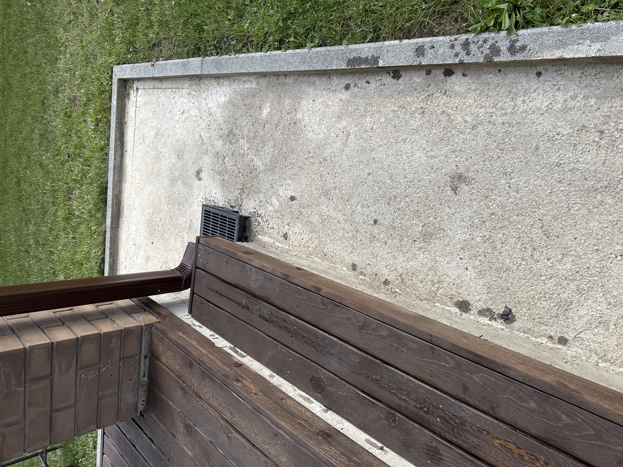 The image shows a drain grate on a concrete patio next to a wooden deck. A downspout from a brown gutter runs down the side of a brick pillar. The patio is bordered by a granite curb and a patch of green grass. There are dark stains on the concrete and the wooden deck appears wet. The scene is outdoors, likely in a residential setting. No people are visible in the image. The location is identified as Дёмино, Russia.