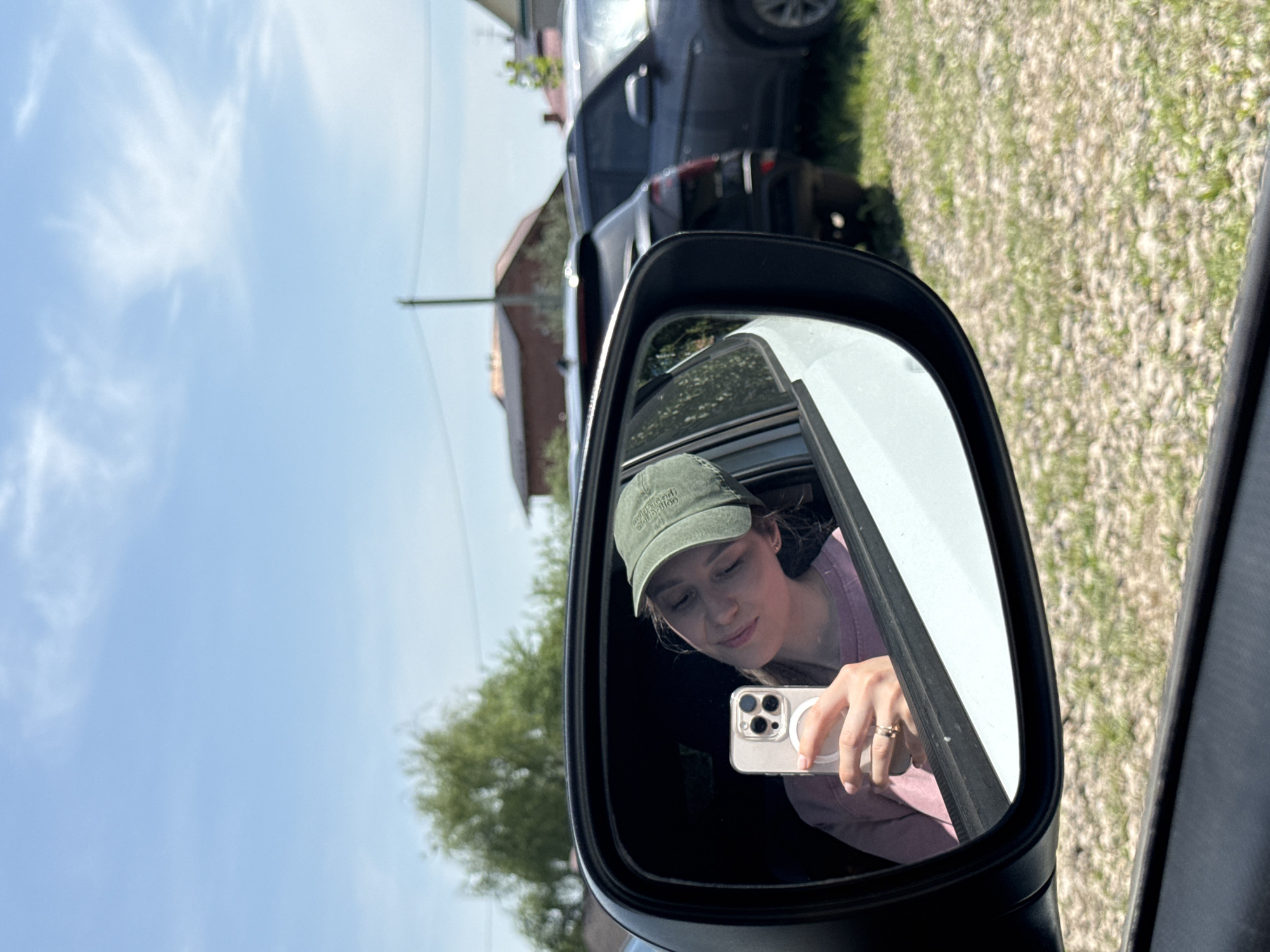 A photograph taken in Прикамский, Russia, shows a woman reflected in a car's side mirror. The woman is wearing a green cap and a purple top. She is holding a white smartphone and taking a picture of herself in the mirror. The reflection shows a glimpse of trees and a building. Outside the mirror, a blue sky is visible. A black vehicle is partially visible in the upper part of the frame, and a gravel surface is seen in the background.