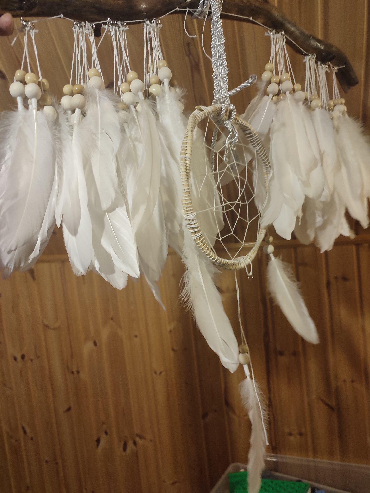 A dreamcatcher made of white feathers and wooden beads hangs from a branch against a wooden wall. The dreamcatcher features a circular woven frame with a web-like pattern inside and is adorned with numerous white feathers suspended from the frame and a branch above. The wooden wall is made of vertical planks with visible knots and grain. The image appears to be taken indoors, likely in a home or workshop, given the presence of what appears to be a plastic storage container in the lower right corner. No people are visible in the image. The location provided is the city of Chashcha, Russia.
