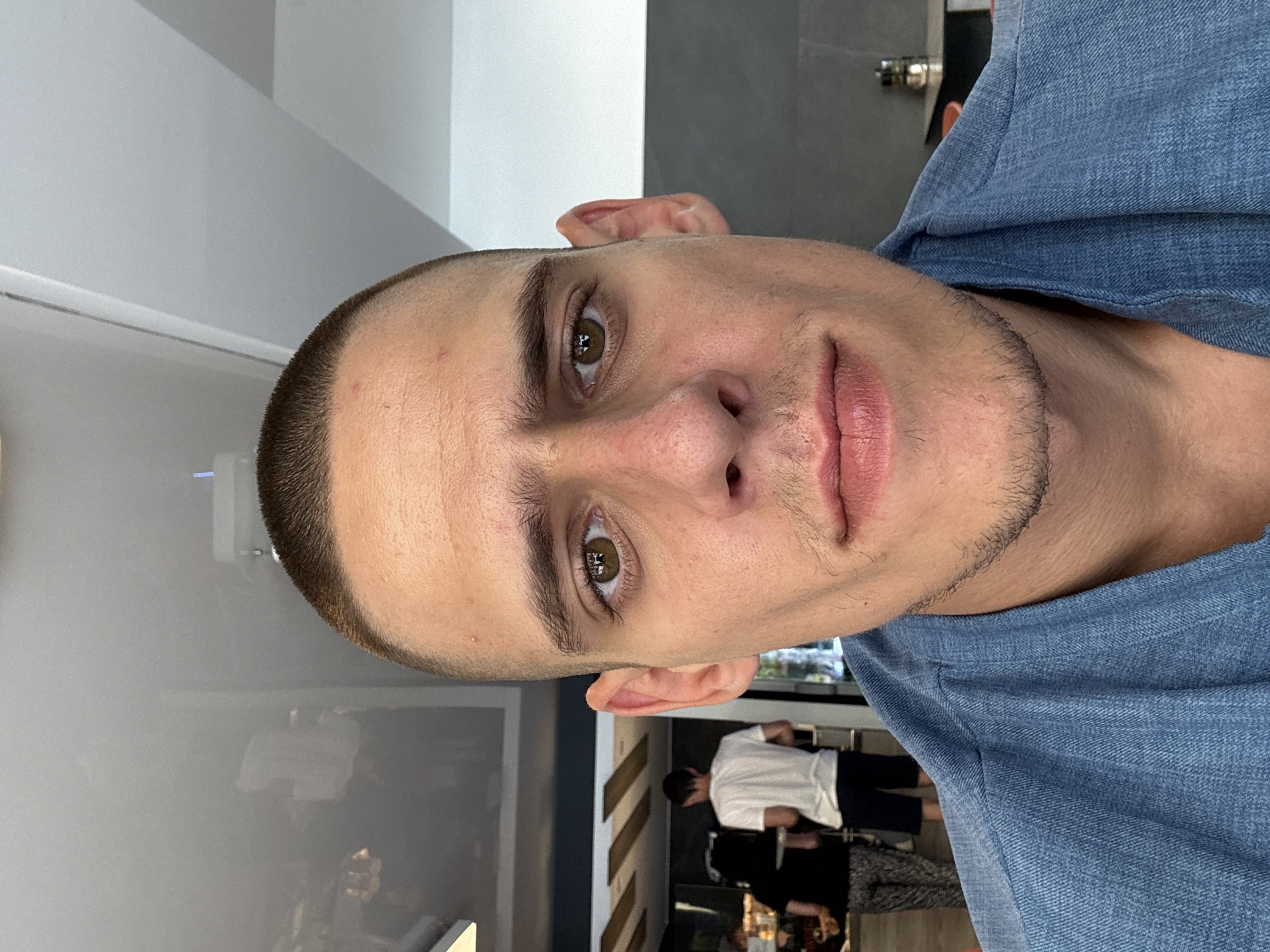 A close-up selfie of a man with a buzz cut, brown eyes, and light stubble. He is wearing a blue denim shirt and is positioned in the foreground. In the background, out of focus, are several people in what appears to be a cafe or restaurant setting, with tables and chairs visible. The overall lighting suggests an indoor environment during the daytime. There is no identifiable information to suggest the location is Sofia, Bulgaria.