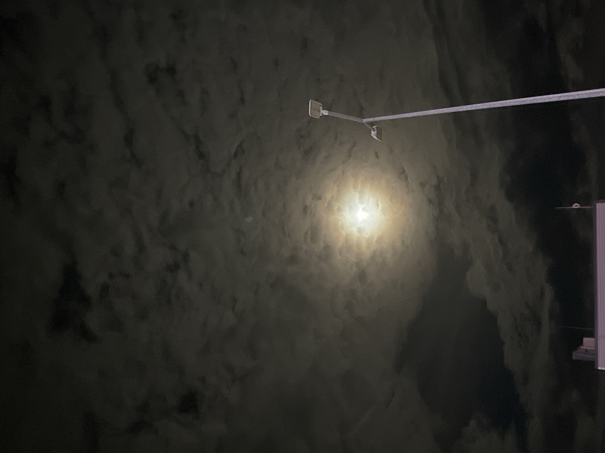 The image captures a night scene in Yambol, Bulgaria, with a street light illuminating a cloudy sky. The moon, obscured by clouds, is visible as a bright, diffused light source. A street lamp with a long, thin arm extends into the frame, its head positioned near the moon. A tall structure, partially visible on the right edge, appears to be the side of a building. The environment is dark, with the clouds adding a textured, somewhat ominous feel to the nightscape. No people or animals are visible.