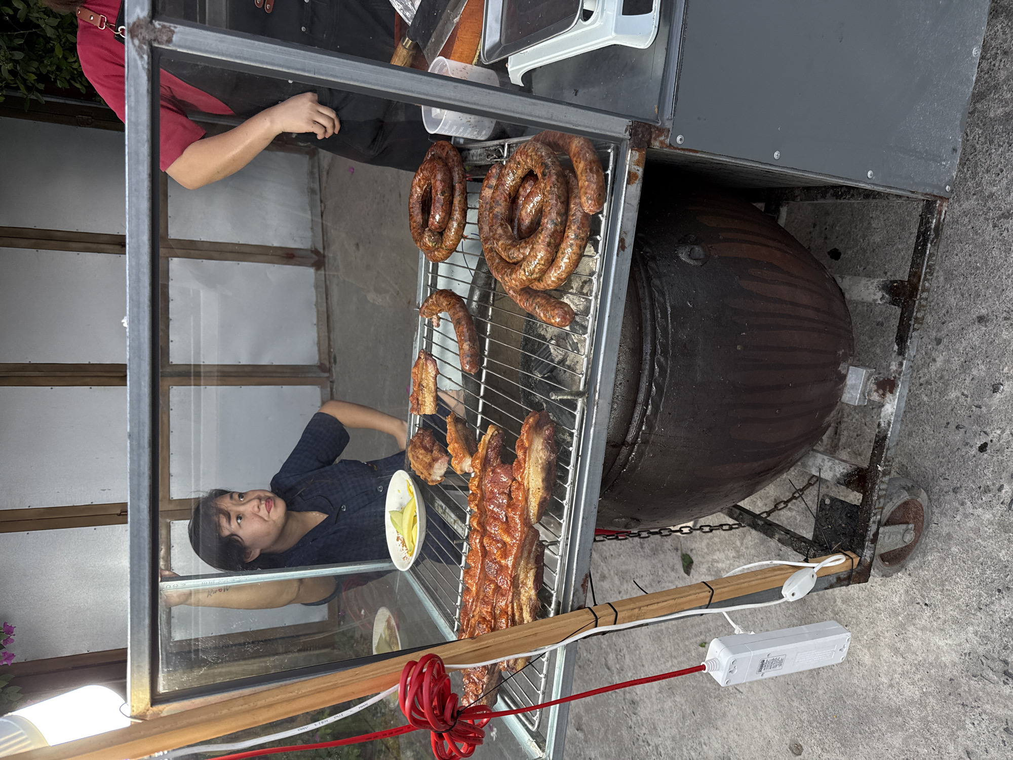 In Bắc Nha Trang, Vietnam, the image depicts a street food vendor within a mobile cart. The vendor, a young Asian individual, is positioned behind a glass display case. The case features a grill laden with an array of cooked meats, including coiled sausages and pork belly. A small dish containing sliced fruit rests near the grill. The cart itself is constructed of metal framing with wood and metal paneling. A large, dark, rounded object, likely a cooking vessel, is situated beneath the grill. Electrical cords and a power strip are visible near the base of the cart. The setting appears to be outdoors, on a paved surface. Another person is visible in the background above the glass display.