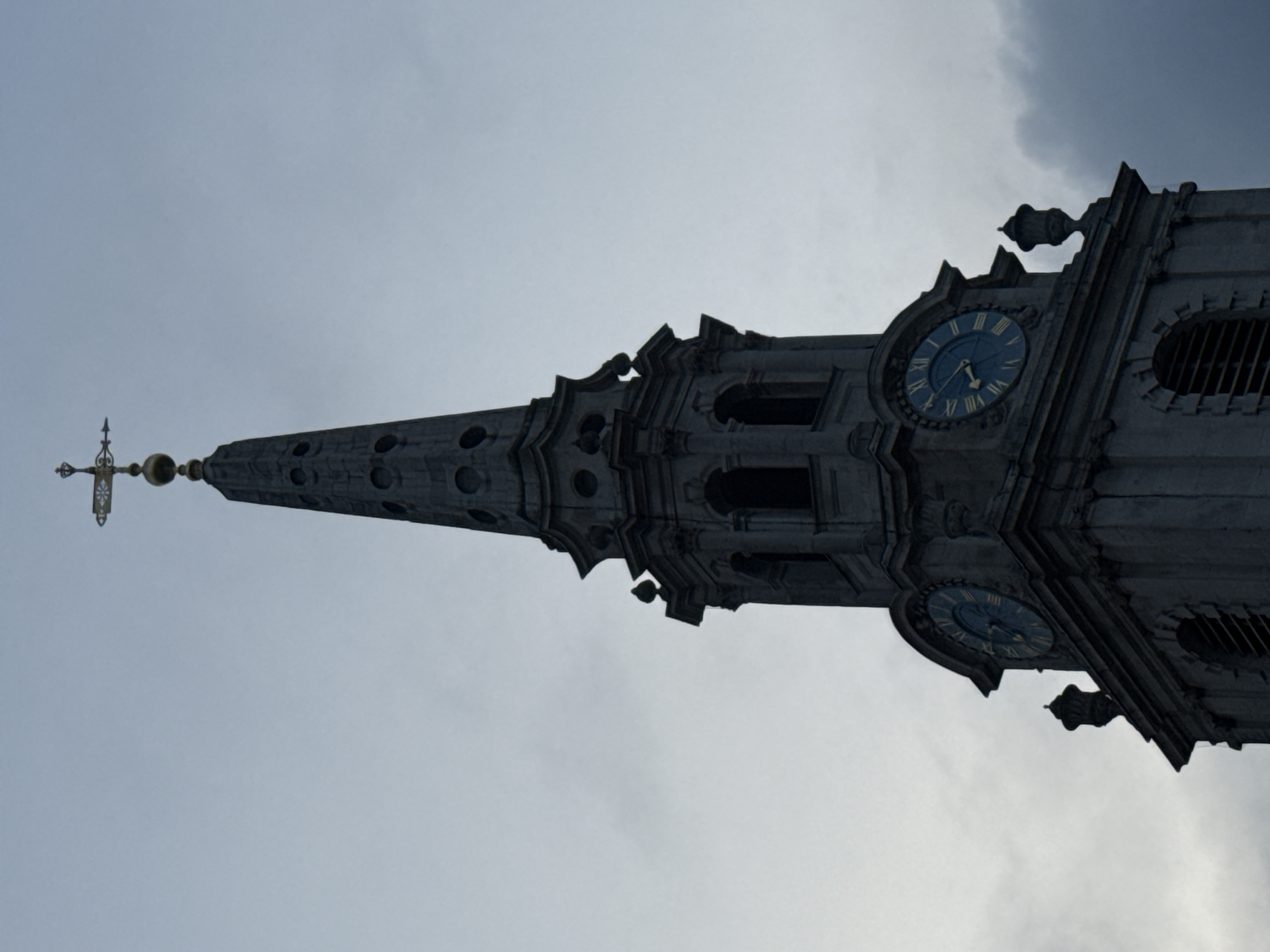 The image shows the upper portion of a tall, ornate clock tower against a cloudy sky. The tower is made of light-colored stone, possibly granite or limestone. It features architectural details such as arched openings, decorative moldings, and a tapering spire adorned with small circular windows or openings. A weather vane or cross tops the spire. Two clock faces are visible, each with Roman numerals and blue clock hands. This is likely the Wren church of St. Vedast Foster Lane in London, UK.