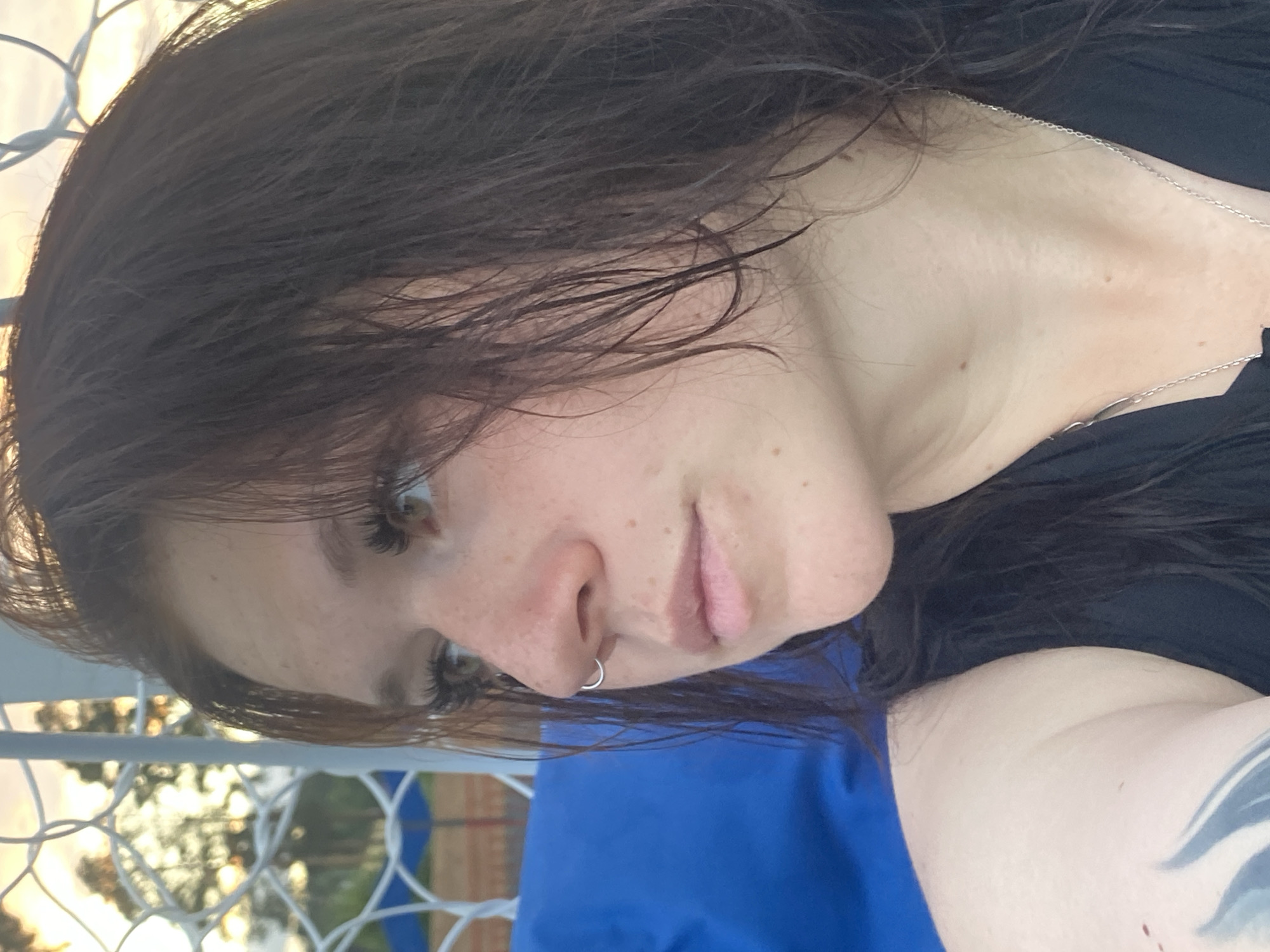A close-up, profile shot captures a young woman with long, dark, wet hair framing her face as she looks to her left. She is wearing a black tank top and a silver necklace. A portion of a blue object, likely a cushion or chair, is visible behind her to the left. In the background, a white fence or netting is present, with trees and the sky visible through it. The lighting suggests it is either sunrise or sunset. There is no indication of the specific location within Votkinsk, Russia.