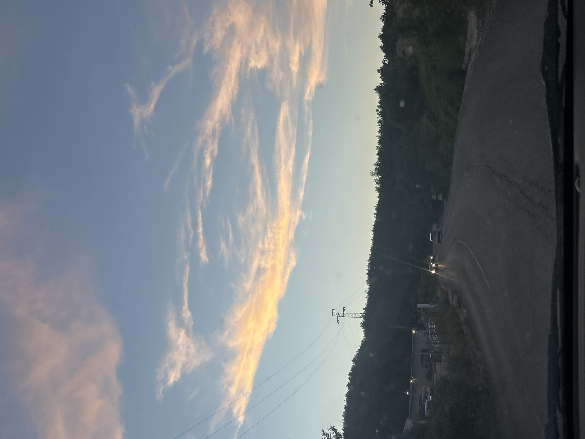 The image depicts a road curving through a wooded, hilly area under a sky filled with wispy, pink-tinged clouds. On the left side of the road, a building with lights is visible, accompanied by a fence and some vehicles. A tall electrical tower with wires stretches across the scene, silhouetted against the sky. Several cars are driving on the road ahead, their headlights illuminated. The overall impression is of a rural or semi-rural setting during dusk or dawn.