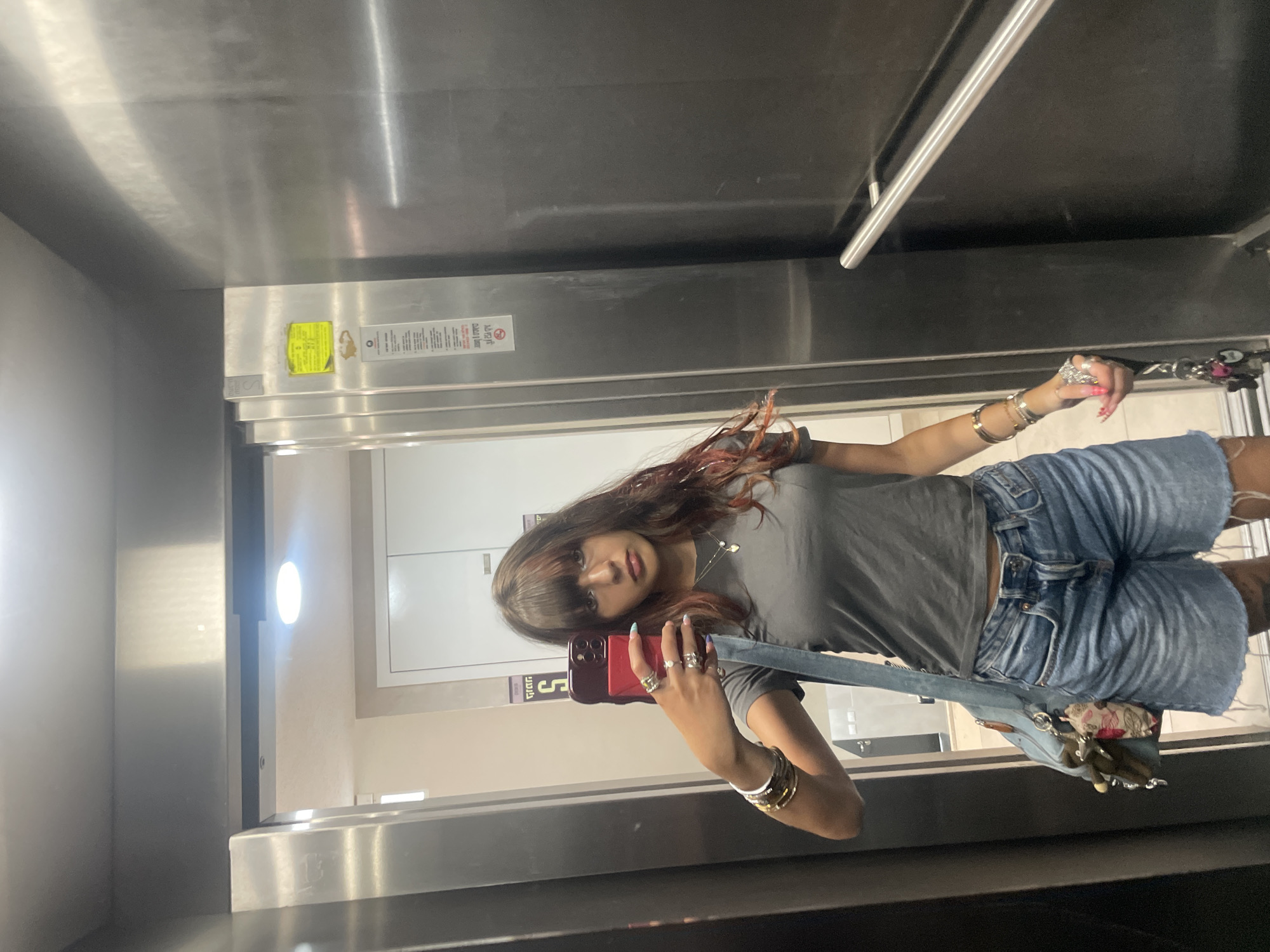 A young woman with long, dark hair and bangs is taking a mirror selfie in an elevator. She is wearing a grey t-shirt, denim shorts, and multiple rings and bracelets. She has a light blue shoulder bag slung across her body. The elevator appears to be modern with stainless steel walls and a silver handrail. In the reflection of the elevator doors, a sticker with Hebrew writing and a yellow sticker with some writing are visible on the wall. This elevator is located in Raanana, Israel.