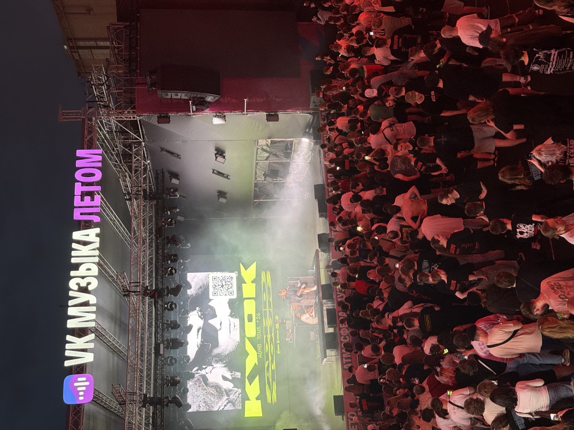 The image depicts an outdoor concert venue at night, likely in Moscow, Russia, given the prominent "VK МУЗЫКА ЛЕТОМ" signage, a well-known Russian social media platform. A large stage is set up with a massive screen displaying a black and white image of a person wearing sunglasses, along with the text "KYOK ADHD TOUR '26" in bright yellow. Two individuals are performing on the stage, one seated and the other standing, with musical equipment between them. A dense crowd of spectators, dressed in casual attire, is gathered in front of the stage, many holding up their phones to capture the performance. The atmosphere is illuminated by stage lighting and the glow of the screen, with smoke effects adding to the ambiance. The overall scene suggests a lively and engaging music event.