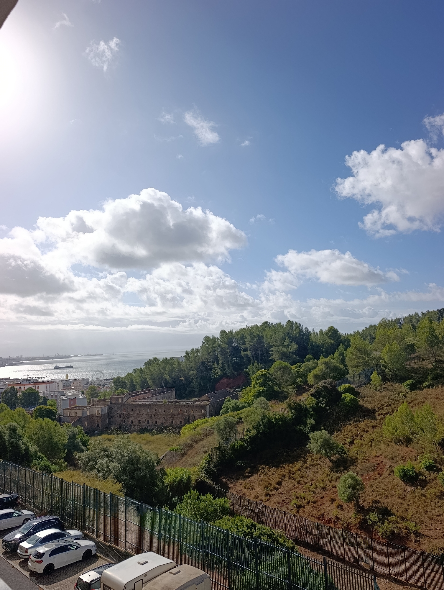 The image depicts a sunlit scene in Setúbal, Portugal, with a view overlooking the city, a port, and a body of water. In the foreground, a row of parked vehicles including cars and a caravan is visible behind a green metal fence. Beyond the fence, a sloping hillside is covered with dry grass and scattered trees. In the middle ground, a weathered stone building, possibly an old fort or industrial structure, sits on the hillside amidst lush greenery. Further into the distance, a cityscape with buildings and a Ferris wheel can be seen along the waterfront, with ships and the expanse of the sea under a bright, cloudy sky. The overall impression is one of a coastal urban environment with historical elements set against a natural backdrop.