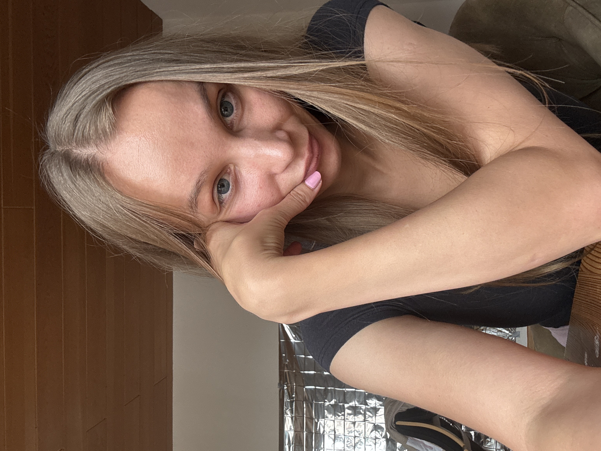 A young woman with blonde hair and blue eyes is looking at the camera with her head tilted and her hand resting on her cheek. She is wearing a dark t-shirt. The background is blurred, but appears to be indoors with wooden paneling on the ceiling and a reflective surface on the wall behind her. There is no information available that would allow for the determination of the image's location in Überherrn, Germany.