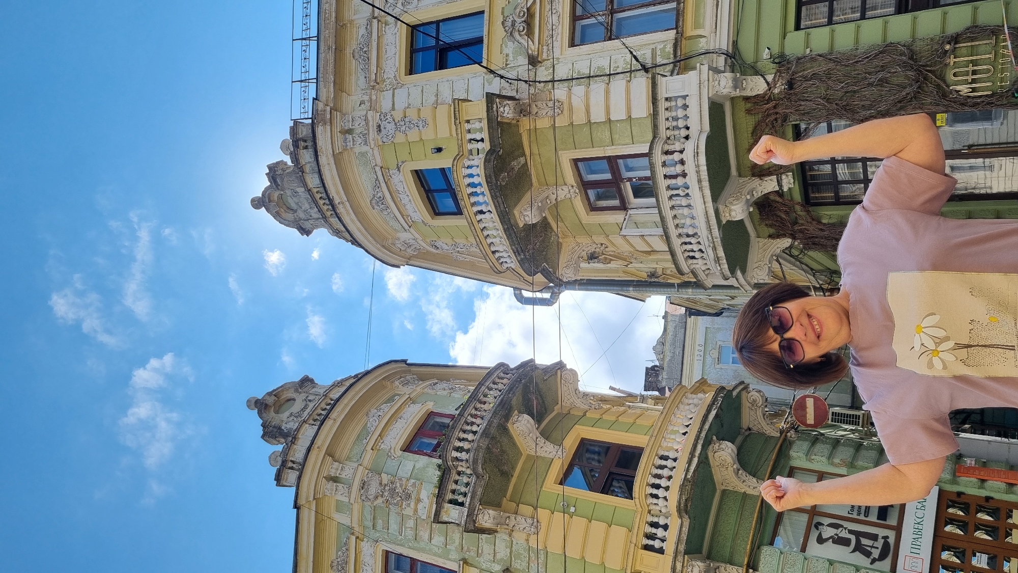 A woman with short brown hair and sunglasses is standing in front of two ornate, pastel-colored buildings with balconies in Chernivtsi, Ukraine. She is wearing a light purple t-shirt with a floral design and is flexing her arms. The buildings are illuminated by sunlight, and the sky is blue with scattered clouds. The buildings appear to be old and have decorative architectural details. A sign on the left building reads "ПРАВЕКС БАНК". The overall impression is that of a sunny day in a historic European city.