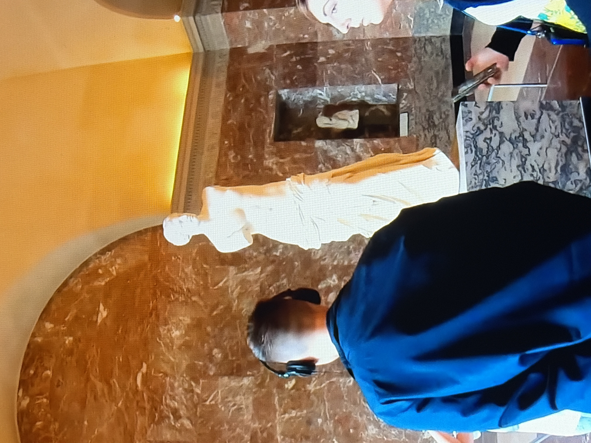 In a museum, a man wearing headphones stands with his back to the camera, facing a statue. The statue, a classical marble figure, is positioned on a pedestal. The walls of the museum are adorned with patterned marble. To the right of the statue, a woman is partially visible, looking towards the statue. The ceiling is arched and illuminated by warm lighting. Another smaller statue is visible in a recessed niche on the wall behind the main statue.