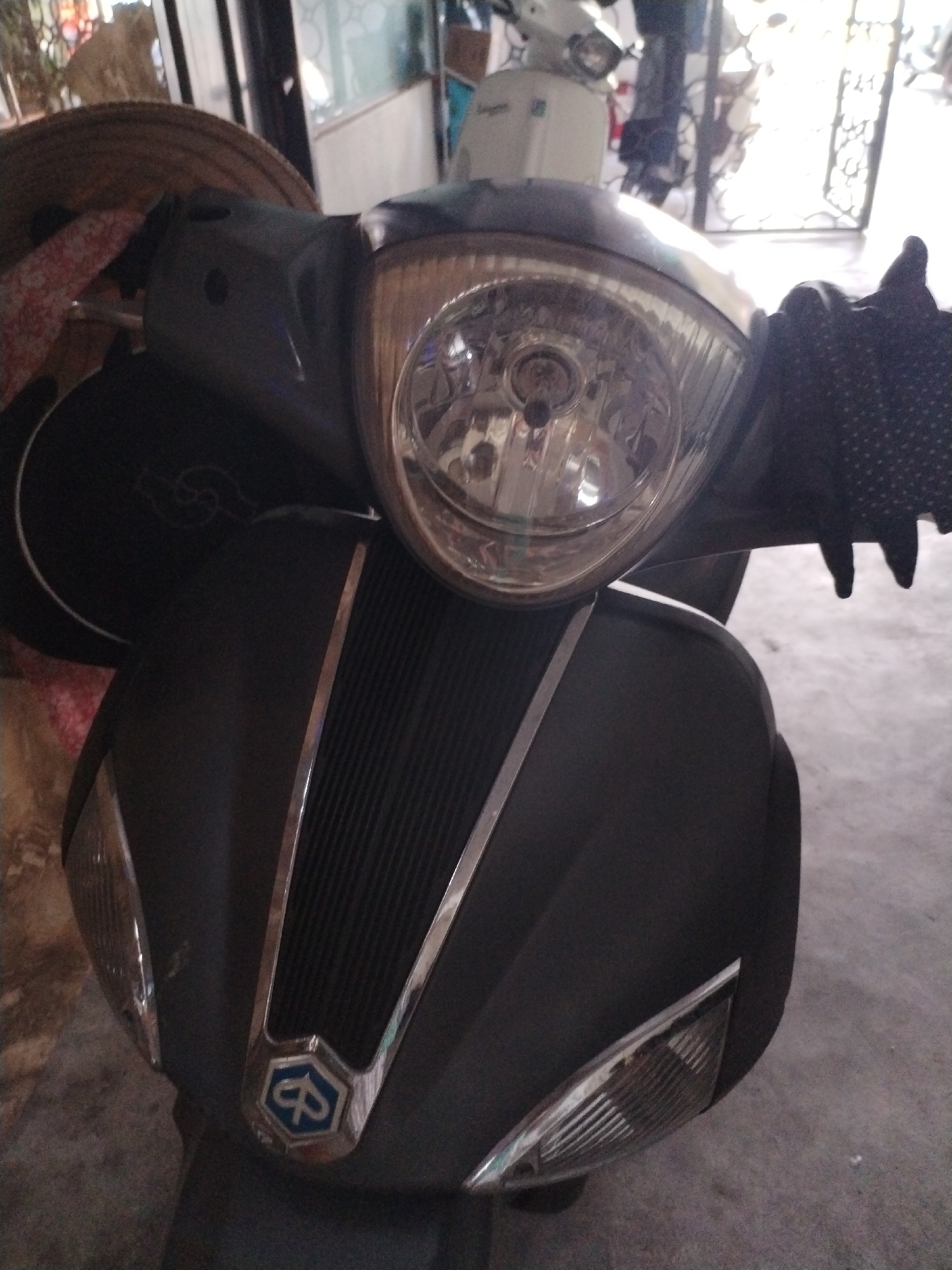 A close-up photograph depicts the front of a dark-colored Vespa scooter. The headlight is prominently featured, along with the distinctive Vespa logo on the front panel. A gloved hand is visible on the right side of the frame. In the background, another white scooter can be seen, partially obscured. The location is in Bac Nha Trang Ward, Vietnam.