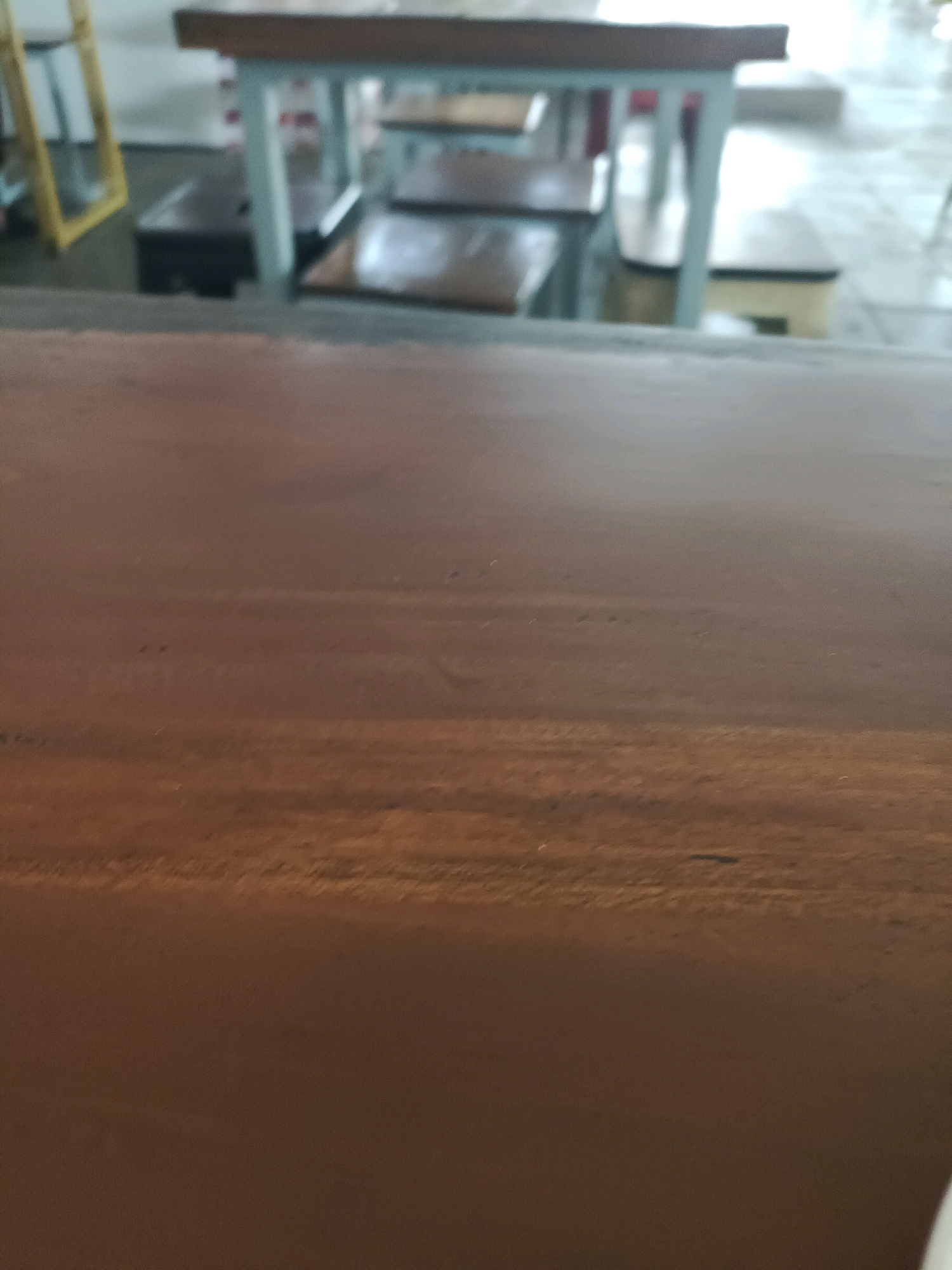 The image depicts a close-up view of a brown wooden surface in the foreground, likely a table or counter. In the blurred background, a light-colored table with a wooden top and light blue legs is visible, with several dark-colored benches or stools placed underneath. The environment appears to be an indoor setting, possibly a cafe or restaurant. The overall lighting is subdued. There are no people or specific actions captured in the frame. The scene does not contain any recognizable landmarks or features that would indicate a specific location in the city of Made, Indonesia.