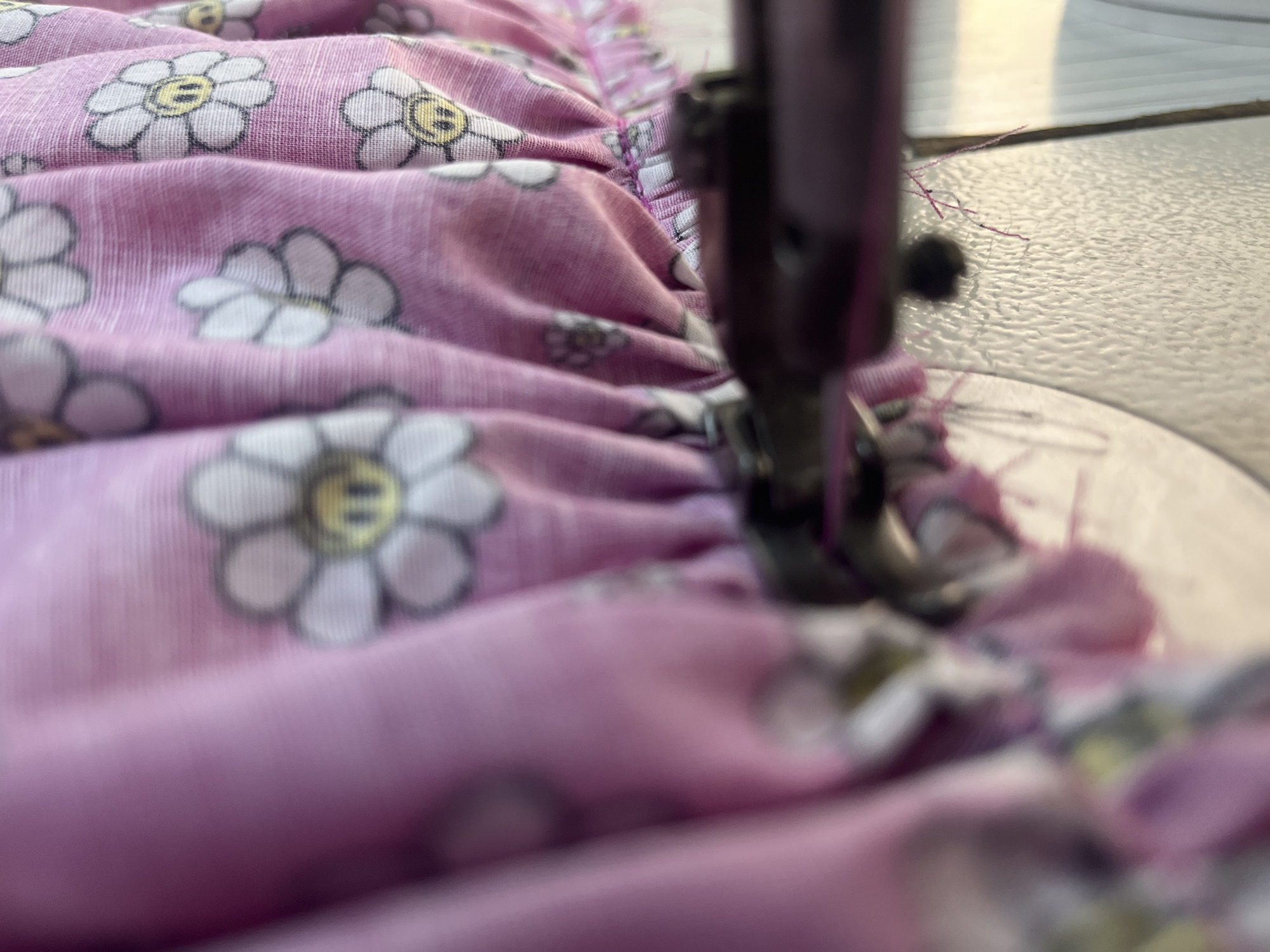 A close-up, slightly blurred, indoor shot shows a pink fabric with a repeating pattern of white daisies with yellow centers being sewn by a sewing machine. The fabric is gathered and pleated. The sewing machine's needle and presser foot are visible in the foreground, out of focus, with pink threads hanging. The background is a blurred light-colored surface, possibly a table.