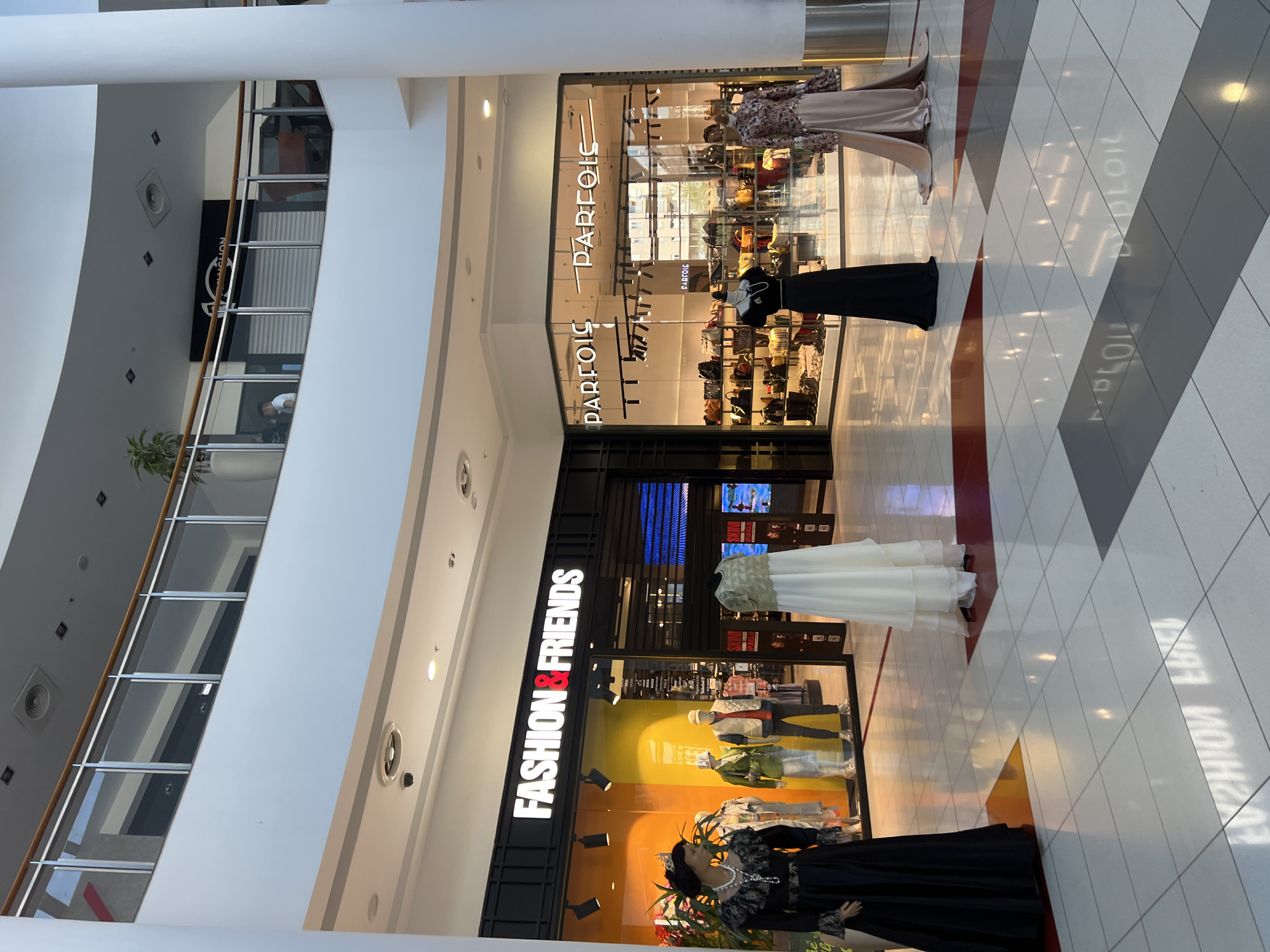 The image depicts the interior of a shopping mall in Podgorica, Montenegro. Two storefronts are prominently featured: "FASHION & FRIENDS" and "PARFOIS." Mannequins dressed in various outfits stand in front of both stores. The mall's architecture includes multiple levels with glass railings and white pillars. The floor is tiled in a geometric pattern of white, gray, and red. The lighting is bright, reflecting off the polished surfaces. The scene is static, with no visible people actively engaging within the frame other than the shop attendants in the stores.