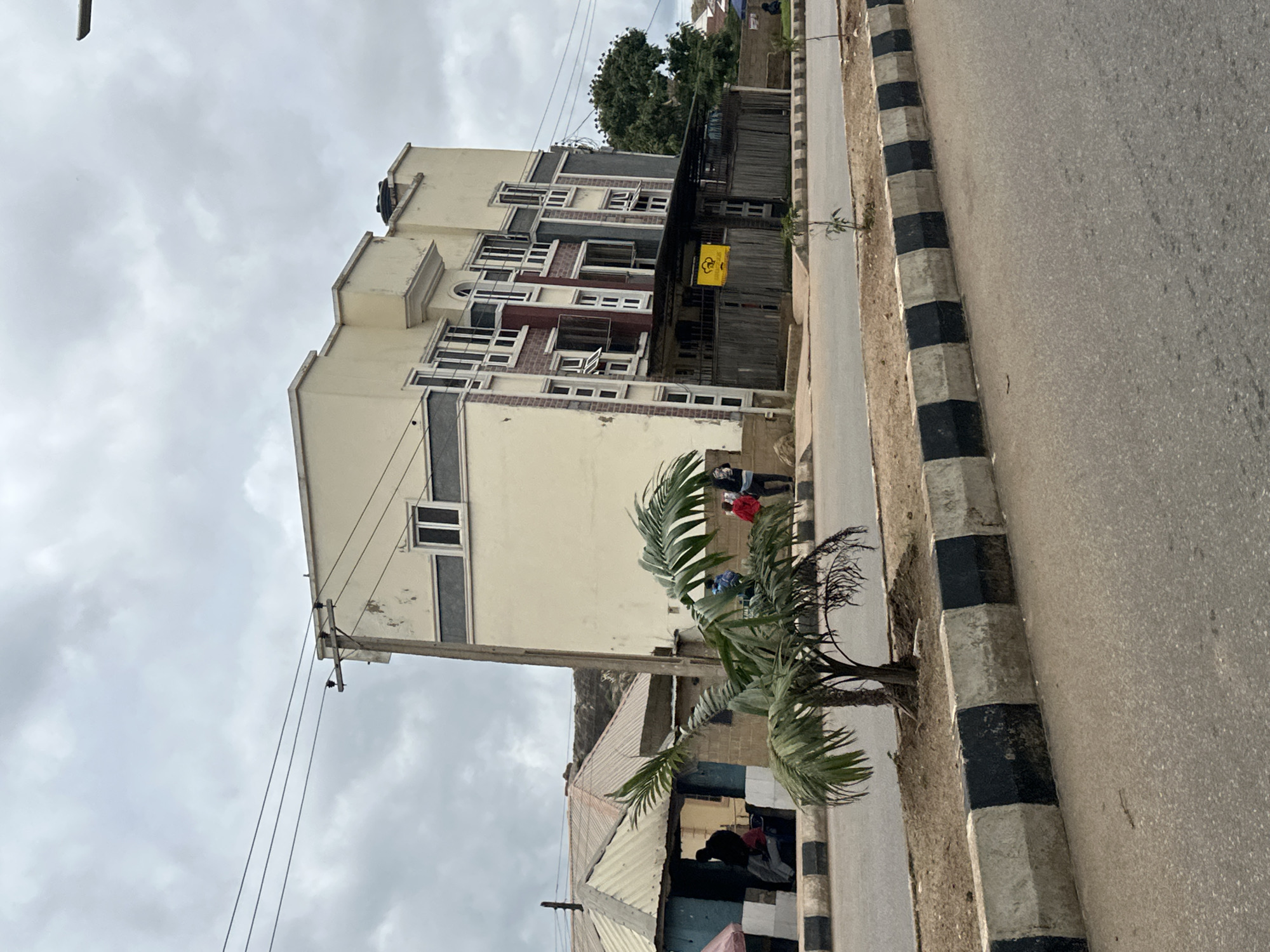 The image depicts a street scene in Gura Top, Nigeria, under an overcast sky. A multi-story building with a cream-colored facade and dark gray accents dominates the right side of the frame. The building features numerous windows, some of which are open. A corrugated metal roof structure, painted in shades of brown and white, is visible to the left of the multi-story building. In front of this structure, a small, open-air stall with blue walls and white trim is occupied by several individuals, some of whom appear to be seated. A young palm tree with green fronds stands in the middle ground, situated between the road and the buildings. Power lines are strung across the top of the image, supported by a concrete pole. The road in the foreground is paved, bordered by a concrete barrier with alternating black and white markings. Several trees are visible in the background, and some individuals are standing near the multi-story building and the smaller stall. A yellow sign with a black illustration is affixed to the fence in front of the multi-story building.