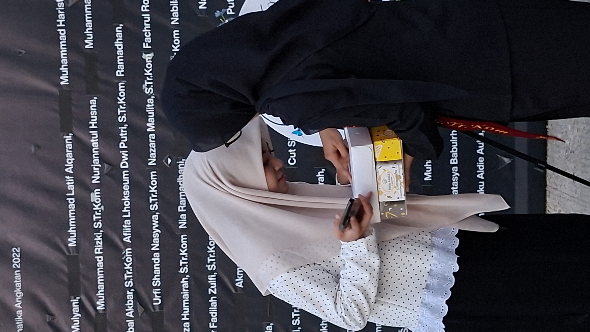 Two women wearing hijabs are interacting in front of a dark banner with white text. The woman on the left, wearing a light-colored hijab and a white polka-dotted shirt with lace trim, is holding a smartphone and looking down at two gift boxes she is holding. The woman on the right, wearing a black hijab, is reaching towards the gift boxes. The banner behind them contains text that appears to be names and titles, with some text readable as "MATIKA Angkatan 2022," and other names like "Mulyani," "Muhammad Latif Alqarani," "Nurjannatul Husna," and possibly locations or institutions related to Lhokseumawe, Indonesia. The environment appears to be outdoors, with the lighting suggesting daytime. The overall scene depicts a presentation or gift-giving event.