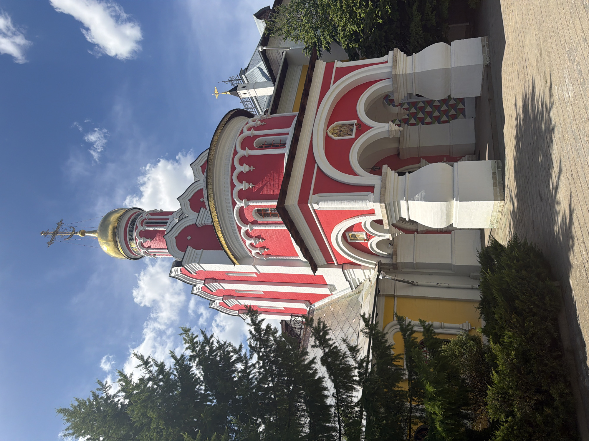 This media file captures a vibrant Russian Orthodox church in Pavlovskaya Sloboda, Russia, under a clear blue sky with scattered white clouds. The church dominates the frame with its striking red and white facade, adorned with intricate architectural details including arched windows, decorative cornices, and small columns. A prominent golden onion dome, topped with a golden Orthodox cross, gleams under the sunlight. Around the base of the dome, there appears to be illegible Cyrillic text.
The lower section of the church on the right features a distinctive geometric pattern of red, green, and blue triangles. Further along the facade, there are small religious icons or images set within arched niches. Part of the church's lower exterior is painted yellow.
In the foreground and surrounding the church are numerous green coniferous trees, adding a natural element to the scene. Behind the main church, a smaller, lighter-colored building with a gray roof and a decorative spire, possibly featuring a golden weather vane shaped like a bird, is partially visible. The ground is paved with what appears to be stone or brick pavers, and a distinct shadow of a tree is cast across it, indicating bright daylight. The overall impression is one of a sunny day with fair weather. No human activity or visible individuals are present in the image.