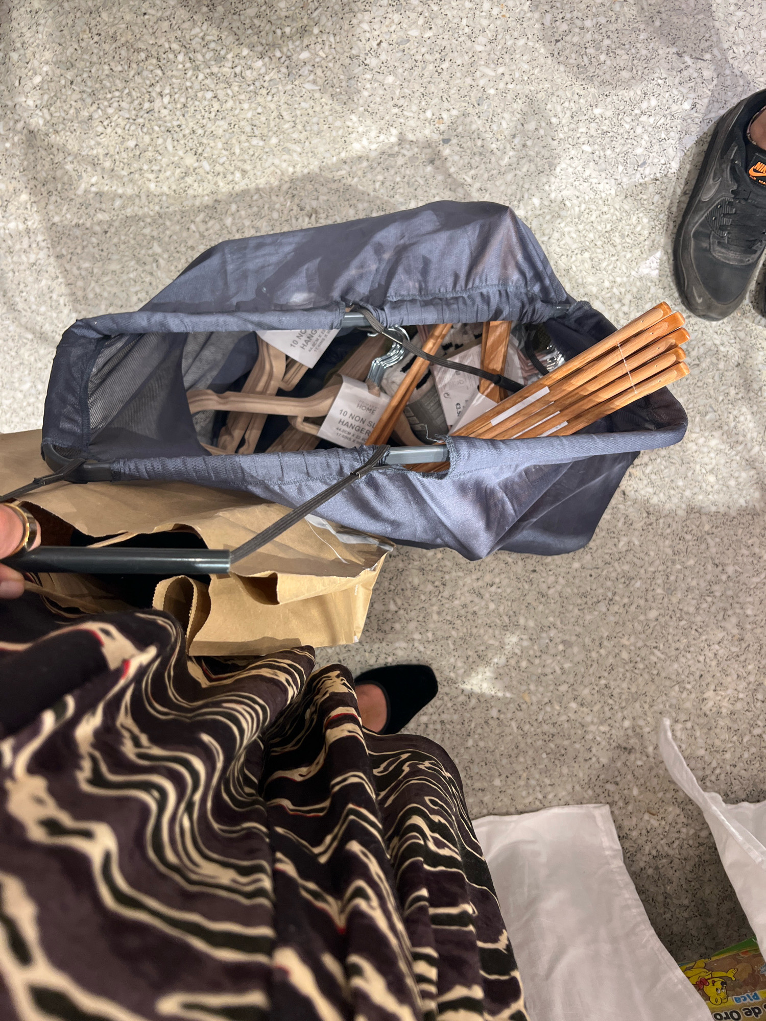In Madrid, Spain, a person's legs and feet are visible, wearing a patterned dress and black shoes. They are standing next to a grey mesh laundry basket overflowing with wooden and plastic clothes hangers. The basket rests on top of brown paper shopping bags, which are on a speckled grey floor. A black Nike Air Max sneaker is also visible on the floor. The scene appears to be indoors, possibly a shopping area or a home.