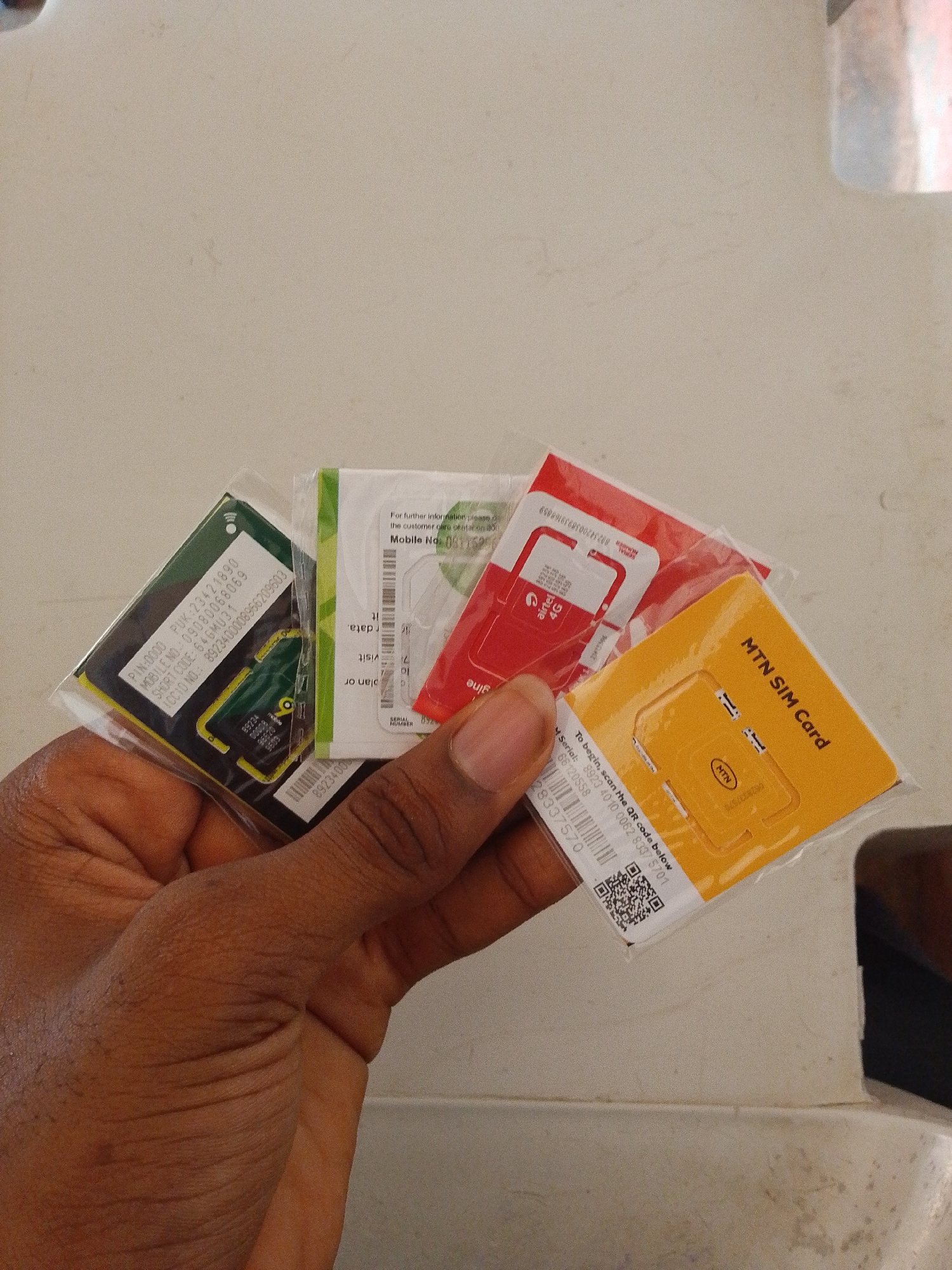 The image shows a person's hand holding multiple SIM cards. The SIM cards are in their original packaging and appear to be from different telecommunication providers, including MTN and Airtel. One card is green, another is red, and a third is yellow with "MTN SIM Card" printed on it. Text on the packaging includes information like PUK codes, mobile numbers, and shop codes. A QR code is also visible on one of the SIM card packages. The background is a plain, light-colored surface, possibly a wall or table, with some subtle texture and faint markings. The lighting suggests an indoor environment. The exact location within Lagos, Nigeria, cannot be determined from the provided image alone.