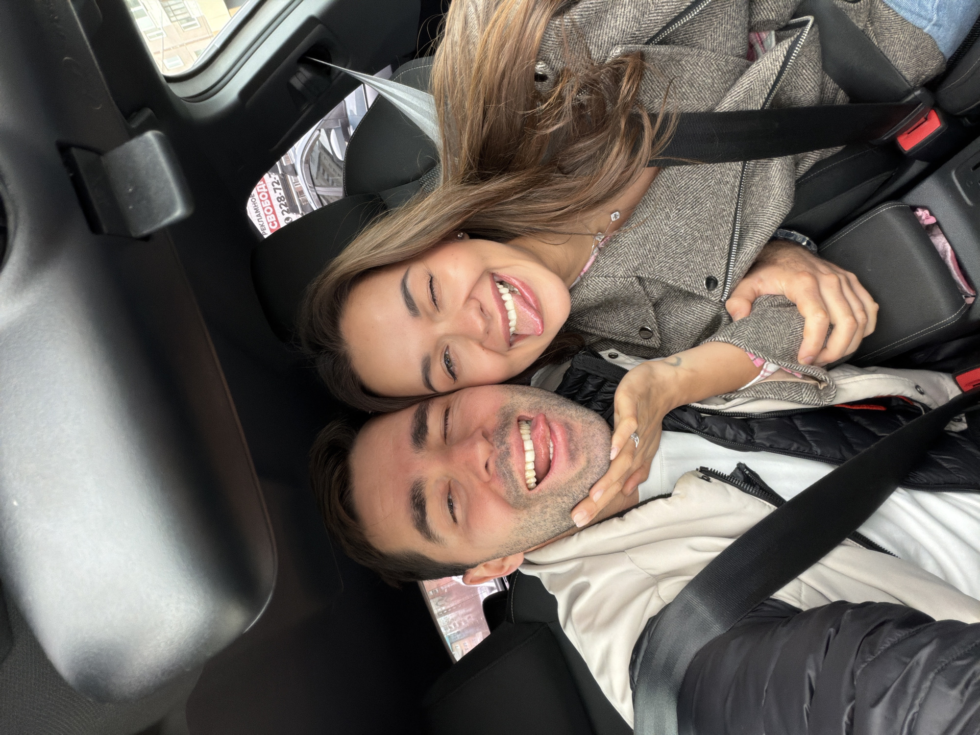Two people are seated in a car. The individuals, a male and female, are wearing seatbelts. The female is wearing a tweed-like jacket with her hair partially covering her face. The male is wearing a light colored jacket over a black puffer jacket. Both are smiling and making playful faces towards the camera. The location is a car, which appears to be in Krasnoyarsk, Russia, based on the Cyrillic text visible on a sticker.