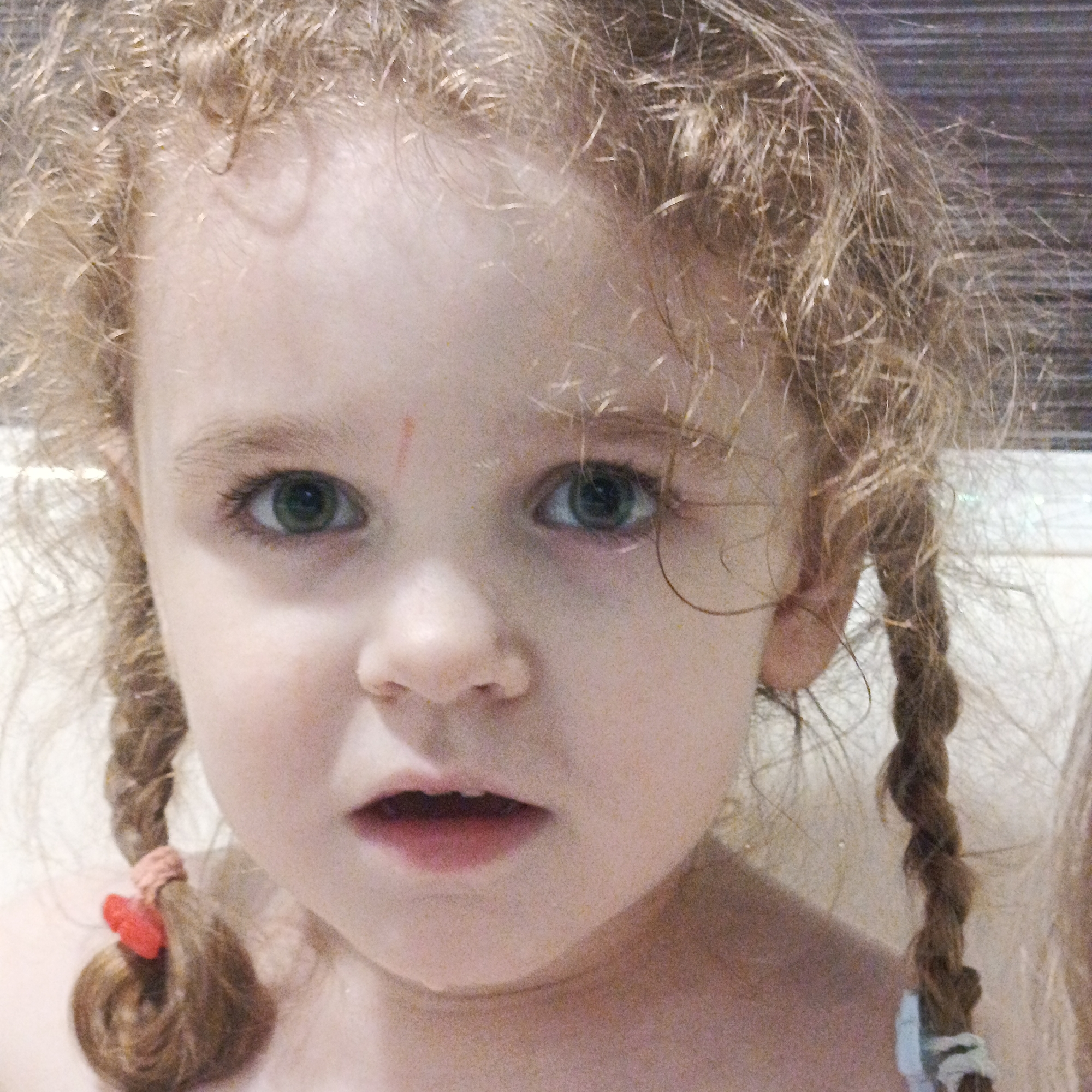 A close-up shot features a young, fair-skinned child with curly, reddish-blonde hair braided into pigtails on either side of their face. Their eyes are a light green color, and their expression is one of slight surprise or uncertainty, with their mouth slightly open. A small, reddish scratch is visible on their forehead. The pigtails are secured with hair ties, one a light pinkish-red with small, stylized red hearts and the other a muted blue. The background is blurred and indistinct. The environment and the city of Марковский, Russia, are not discernible from this image.