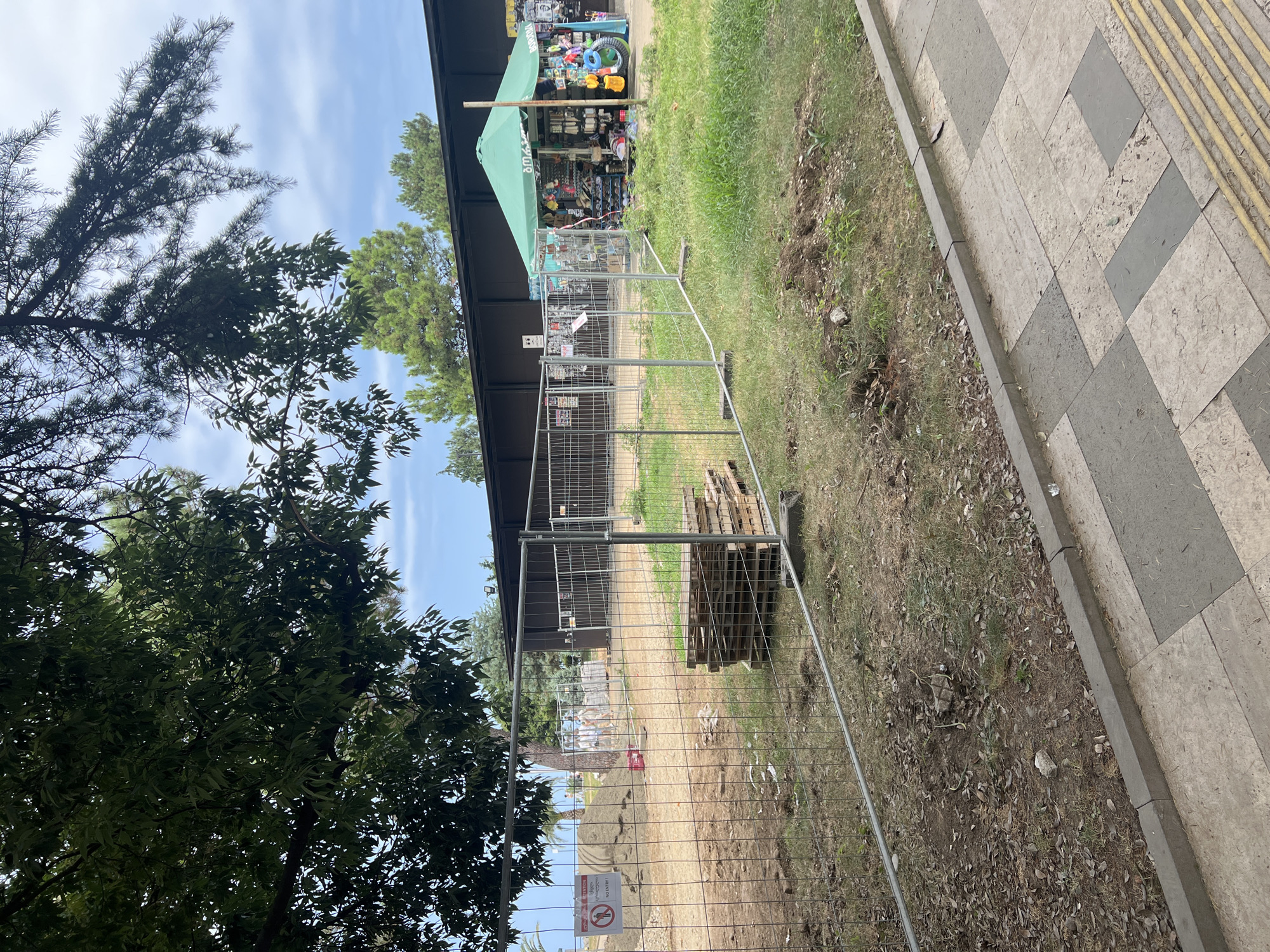 The image depicts a construction site in Batumi, Georgia, cordoned off with temporary fencing. The foreground shows a grassy area with a stack of wooden pallets, and beyond that, a sandy patch with a pile of dirt. A sign reading "ATTENTION" and "NO ENTRY" is visible on the fence. In the background, a small green tent is set up, displaying various items for sale, suggesting it is a temporary market stall. The area appears to be a public space, possibly a beachside location, given the glimpse of the sea and palm trees in the distance. The sky is overcast with scattered clouds.