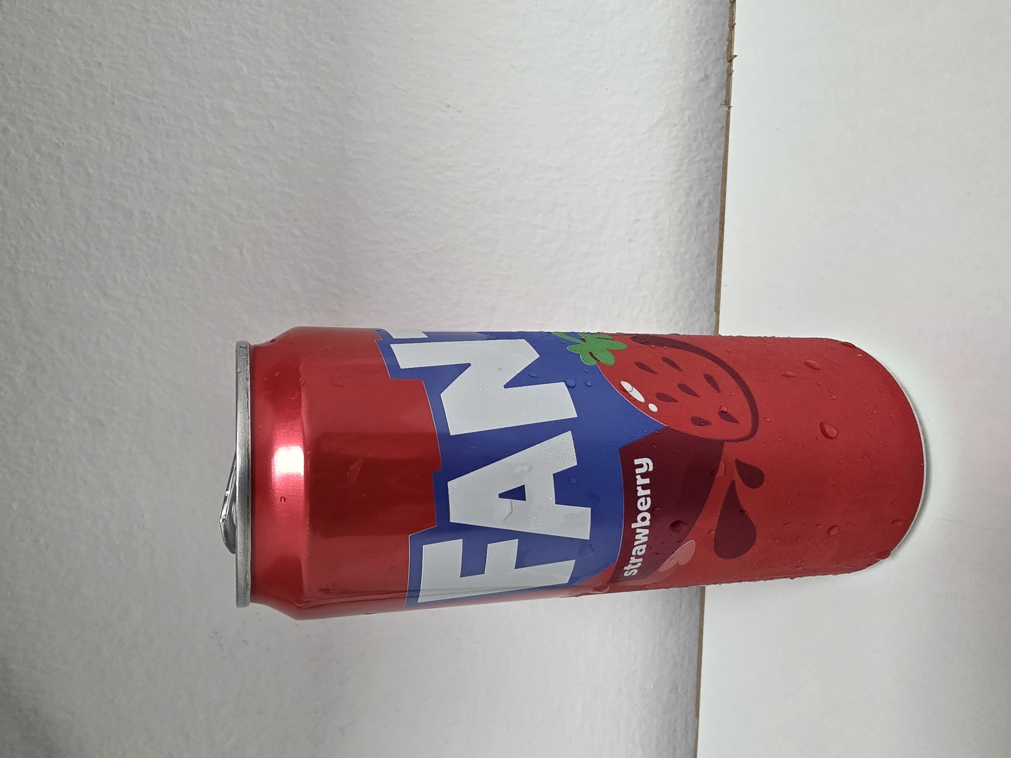 A red Fanta can, frosted with condensation, rests on a white surface. The can features the Fanta logo in blue and white, with the word "strawberry" printed in white text beneath a stylized strawberry graphic. The background is a textured, light-colored wall. The location is not specified in the image, therefore it cannot be identified as Dibba Province, Oman.