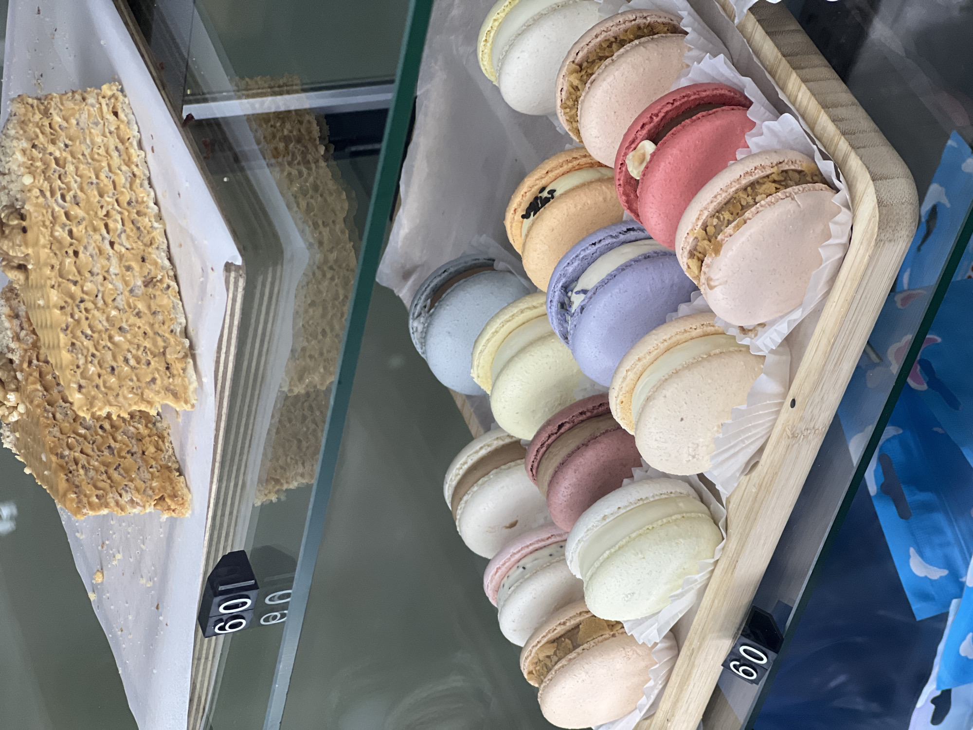 The image displays a vibrant selection of sweet treats in what appears to be a bakery or cafe, viewed through a glass display case.
On the right side of the frame, a wooden tray holds multiple rows of colorful macarons, each placed in a white paper liner. The macarons come in various pastel shades including white, pale yellow, light blue, lavender, light orange, pale pink, and a reddish-brown. Several macarons feature distinct fillings and toppings; for example, one orange macaron is topped with black seeds, while a reddish-brown one has a nut embedded in its shell. Their characteristic "feet" are visible, indicating their delicate structure.
To the left, a large, textured bar of a golden-brown sweet confection rests on white parchment paper. Its surface has a porous, bubbly appearance, suggestive of a crisp nougat, peanut brittle, or a rice crispy-style bar, with visible embedded nuts or grains.
A small black block with the number "60" displayed in white is positioned in the foreground, likely serving as a price tag for the items. Reflections of both the textured bar and the "60" block are visible in the glass behind them. The glass also reflects a blue object with white patterns, possibly a bag or signage, on the far right. The lighting is bright, illuminating the desserts and making their textures and colors stand out.