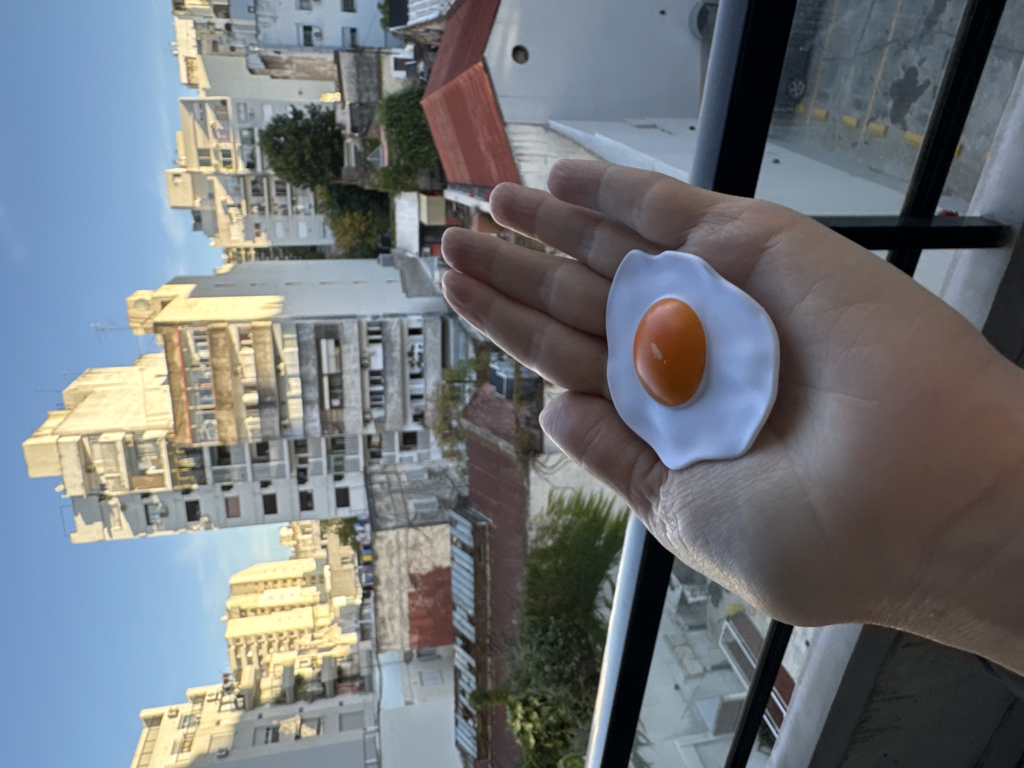 A human hand, light-skinned and positioned in the foreground, holds a small, artificial object designed to look like a fried egg. The "egg" features a white, irregular-shaped "white" surrounding a prominent, spherical orange "yolk." The hand is held out over what appears to be a balcony railing, part of which is visible in the lower right.
The background reveals a bustling urban landscape, a view from an elevated perspective overlooking a dense city. Numerous multi-story buildings, ranging in age and architectural style, fill the frame. Many are concrete or light-colored structures with balconies, some showing signs of wear. Interspersed among the buildings are pockets of green foliage and trees. Distinctive red-tiled roofs are visible on some lower structures. The sky above is a clear, bright blue.
The time of day appears to be late afternoon or early morning, characterized by warm, soft sunlight illuminating the facades of some buildings and casting long shadows, indicating a low sun angle. The weather is clear and sunny. The architectural style and dense urban fabric are consistent with the provided location context of Buenos Aires, Argentina. No discernible text is visible in the image. The scene suggests a moment of playful interaction with the "fried egg" object against an expansive city backdrop.