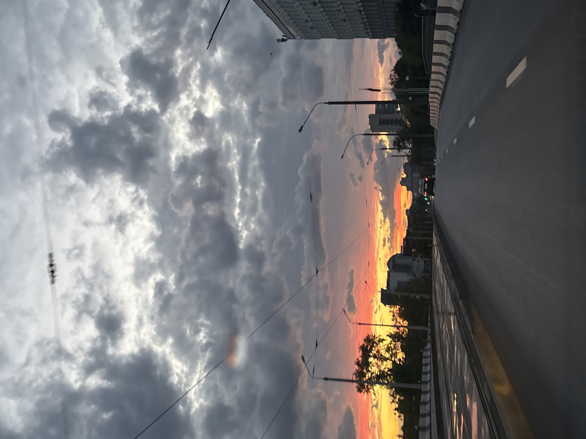 The image shows a view of a street in Minsk, Belarus, at sunset. A wide road stretches across the frame, with a sidewalk or barrier visible on one side. Buildings and trees line the horizon. The sky is filled with dramatic clouds, with hues of orange and red visible near the horizon, indicating the setting sun. Streetlights are positioned along the road.