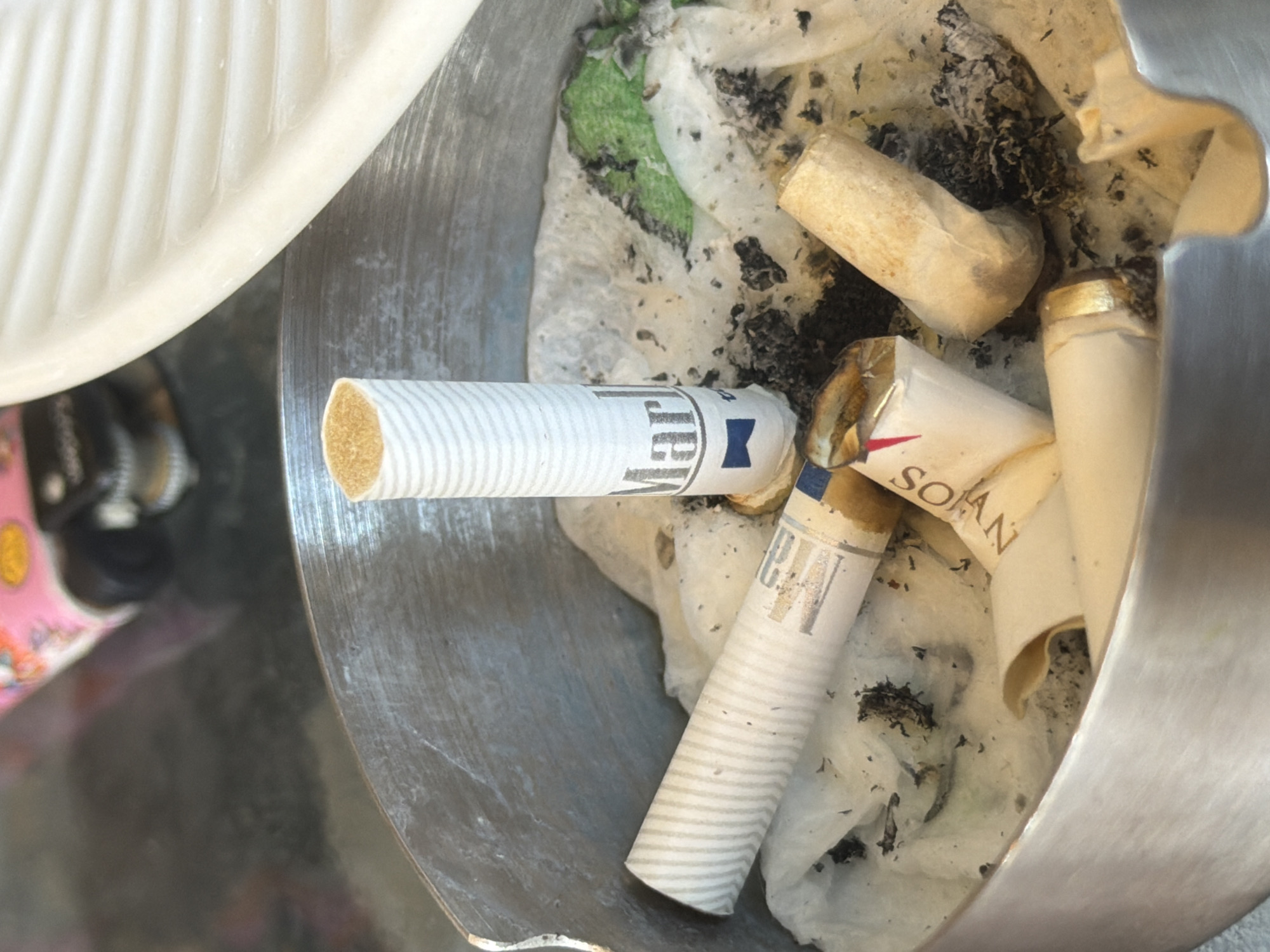 An overhead close-up shot depicts a metal ashtray, containing several cigarette butts amidst ash and debris. The butts show branding from various cigarette brands. Part of a white object is visible in the upper left corner, outside the ashtray.