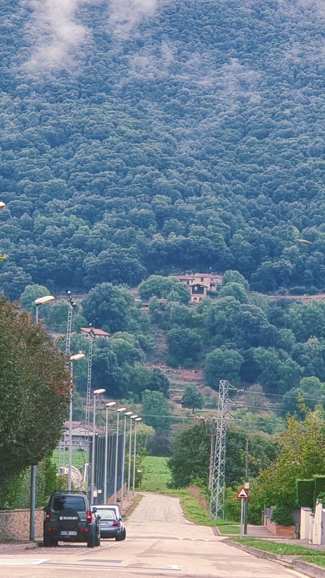 The image depicts a road leading into a densely forested hillside in Sant Joan les Fonts, Spain. In the foreground, a dark green Suzuki SUV is parked on the left side of the road, with a white car parked behind it. A line of streetlights runs along the left side of the road. The road curves upwards and away from the viewer, with a speed bump visible in the asphalt.
Further up the road, a tall electricity pylon stands on the right side, accompanied by a triangular warning sign with a red border and a black graphic. A house with an orange-tiled roof and light-colored walls is visible nestled amongst the trees on the hillside. The background is dominated by a vast expanse of lush, dark green trees, with a misty or cloudy layer obscuring the very top of the mountain. The overall impression is a rural, somewhat remote setting.