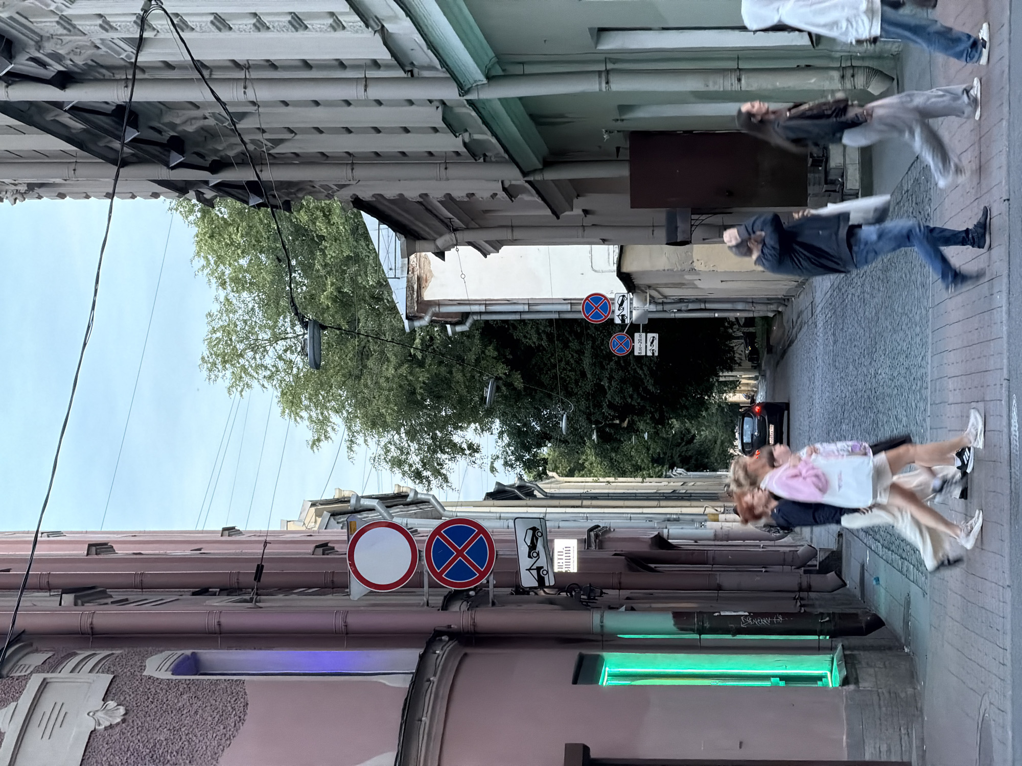 This is a candid, vertical outdoor photograph taken on a street in Saint Petersburg, Russia. The scene is a narrow street lined with buildings on both sides, with trees visible overhead. The perspective is from a low angle, looking upwards and slightly to the right.
The main subjects are several people walking along the sidewalk. Their figures are blurred, suggesting motion and a fast shutter speed or long exposure. One person in the foreground, wearing a black jacket and blue jeans, is walking towards the viewer. Further up the street, a group of three individuals are walking away from the camera, also blurred by movement.
Prominent objects include traffic signs on poles. One sign displays a white circle with a red border, indicating a prohibition. Another sign next to it features a blue "X" within a red circle, signifying a no-parking zone. A third sign shows a silhouette of a car being towed. The architecture of the buildings appears old, with various pipes and drain spouts running along their facades. A green neon light is visible on the lower left side of the frame, emanating from within one of the buildings.
The lighting suggests it is daytime, possibly late afternoon or early evening, given the quality of light and the absence of harsh shadows. The sky is overcast with a pale blue hue. The overall mood is dynamic and urban, capturing a fleeting moment of everyday life on a city street. The blurred motion of the people creates a sense of energy and movement.
There is some text visible on the traffic sign: "8.00-20.00", indicating a time restriction. The architecture and the presence of specific traffic signs could be indicative of a European city, consistent with the provided location context.