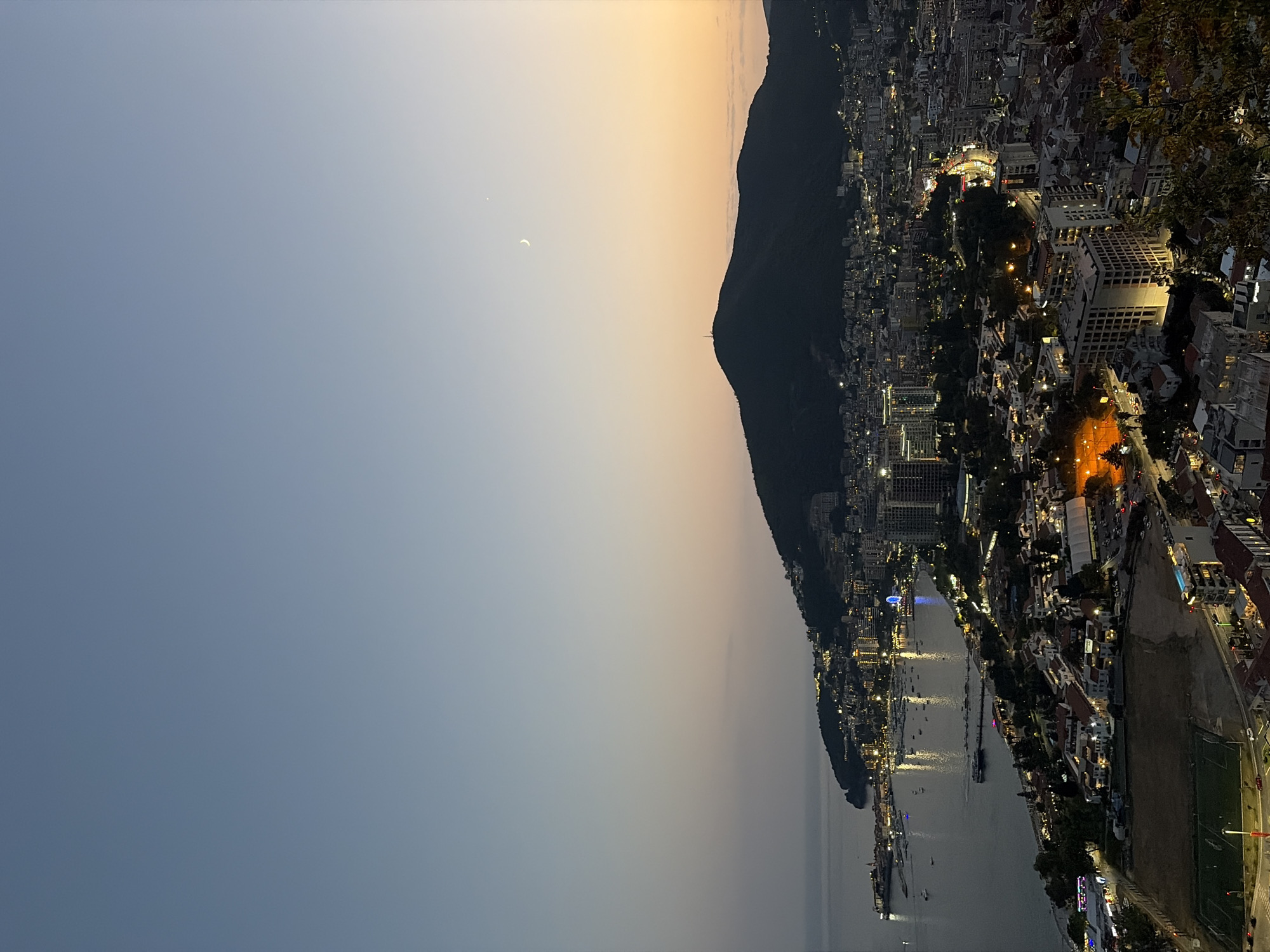 This is a vertical, wide-angle aerial photograph of the coastal city of Košljun, Montenegro, taken at dusk. The sky transitions from a clear, pale blue at the top to a gradient of warm orange and yellow near the horizon, with a faint crescent moon and a bright star visible in the upper right quadrant. The city sprawls across rolling hills that descend towards a dark, tranquil sea. Numerous boats are anchored in the harbor, their lights reflecting on the water. The city itself is illuminated, with buildings, streetlights, and the lights of a Ferris wheel creating a network of glowing points against the darkening landscape. A prominent, dark, tree-covered mountain dominates the mid-ground, with a telecommunications tower visible at its summit. In the foreground, the lights of more densely built areas are visible, including what appears to be a sports field or recreational area. The overall atmosphere is serene and picturesque.