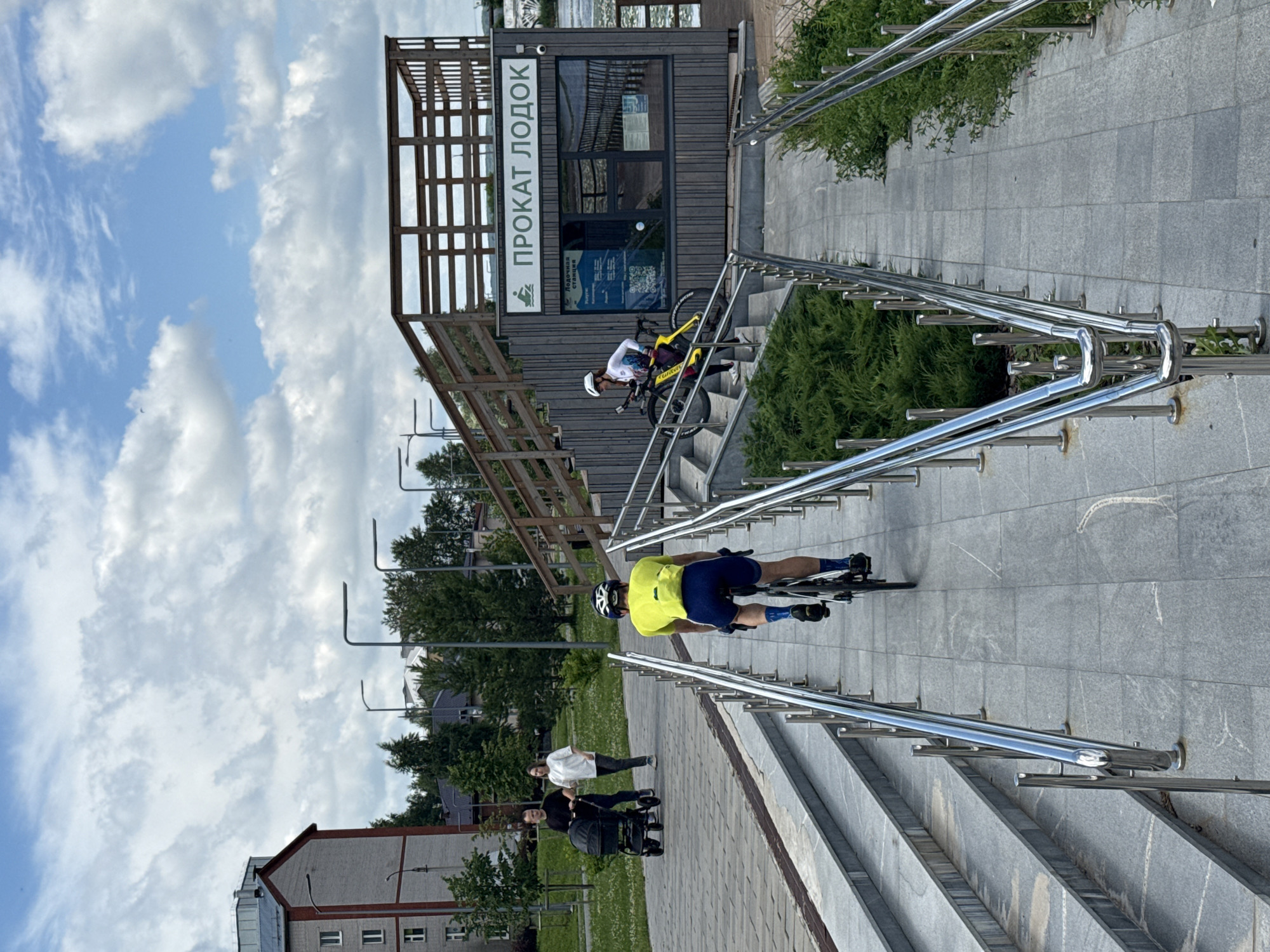 This image depicts a daytime scene in Votkinsk, Russia, specifically along a waterfront promenade. In the foreground, a man wearing a bright yellow shirt and blue cycling shorts is riding a bicycle up a wide, grey stone staircase. He is facing away from the camera and is wearing a helmet. To his left, a woman in white is pushing a stroller with a man in black beside her, also walking on the promenade. Further up the staircase and to the right, another cyclist, a woman in white with a helmet, is seen walking her yellow bicycle up the stairs. Behind the cyclists and people, there is a wooden structure with a sign that reads "ПРОКАТ ЛОДОК" (Boat Rental) in green lettering. This building appears to be a boat rental station. The environment includes green bushes lining the sides of the staircase and promenade, a cloudy blue sky, and in the background, a multi-story building and trees. The overall scene suggests a recreational area in the city.