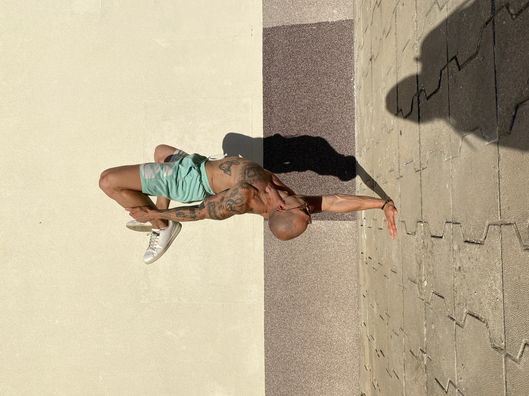 An athletic, bald man with extensive tattoos on his arms and torso performs a handstand outdoors against a textured, two-toned wall. He is wearing light turquoise shorts and white sneakers. His right leg is bent, with his right foot resting on his bent left thigh, while his left leg is extended upwards. His head is tilted downwards, and he wears glasses. The ground beneath him is paved with interlocking stone tiles. A shadow of the man is cast on the ground to his right, and a shadow of his head is visible further to the right. The wall appears to be part of an urban building, possibly in Pleven, Bulgaria, indicated by the architectural style and paved ground. The scene is illuminated by bright sunlight, creating strong shadows.
