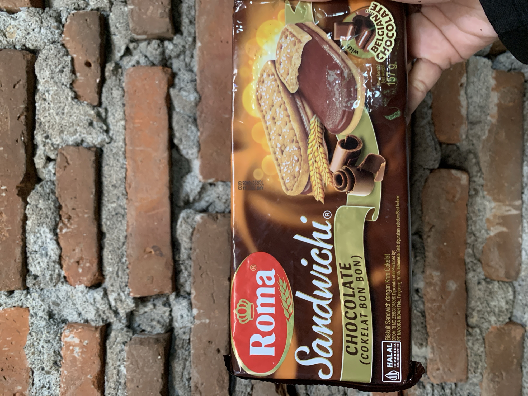 A package of Roma Sandwhichi Chocolate cookies is held in a person's hand against a brick wall. The cookies are chocolate flavored sandwich cookies with a cream filling. The package has "ROMA SANDWICHI CHOCOLATE (COKELAT BON BON)" printed on it. The cookies are depicted with a chocolate filling and a biscuit exterior. There are also chocolate curls and pieces of chocolate shown on the packaging. A "HALAL" certification is visible on the lower left side of the package, along with Indonesian text indicating it is produced by PT MAYORA INDAH Tbk. in Tangerang, Indonesia. The brick wall is composed of reddish-brown bricks with grey mortar. The background is out of focus, but appears to be an outdoor setting. There is no indication of the location in Karangkemiri, Indonesia.