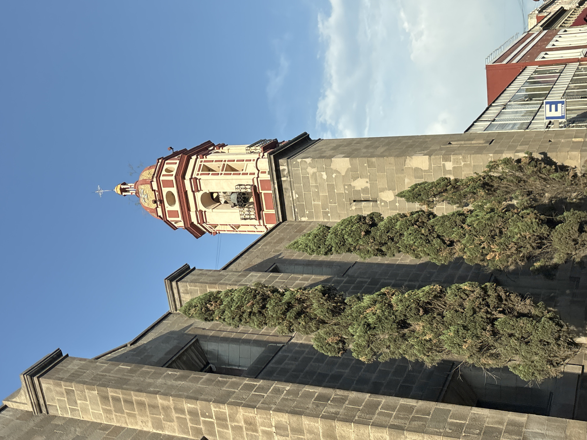 The image shows a tall, ornate church tower against a blue sky with some clouds. The tower is constructed of grey stone blocks and features a bell tower with red and cream-colored trim, topped with a red and gold dome and a cross. Rows of trees are planted along the side of the church building, partially obscuring its features. Adjacent to the church is a modern building with a red roof and glass windows. The environment suggests the location is the city of Puebla, Mexico.