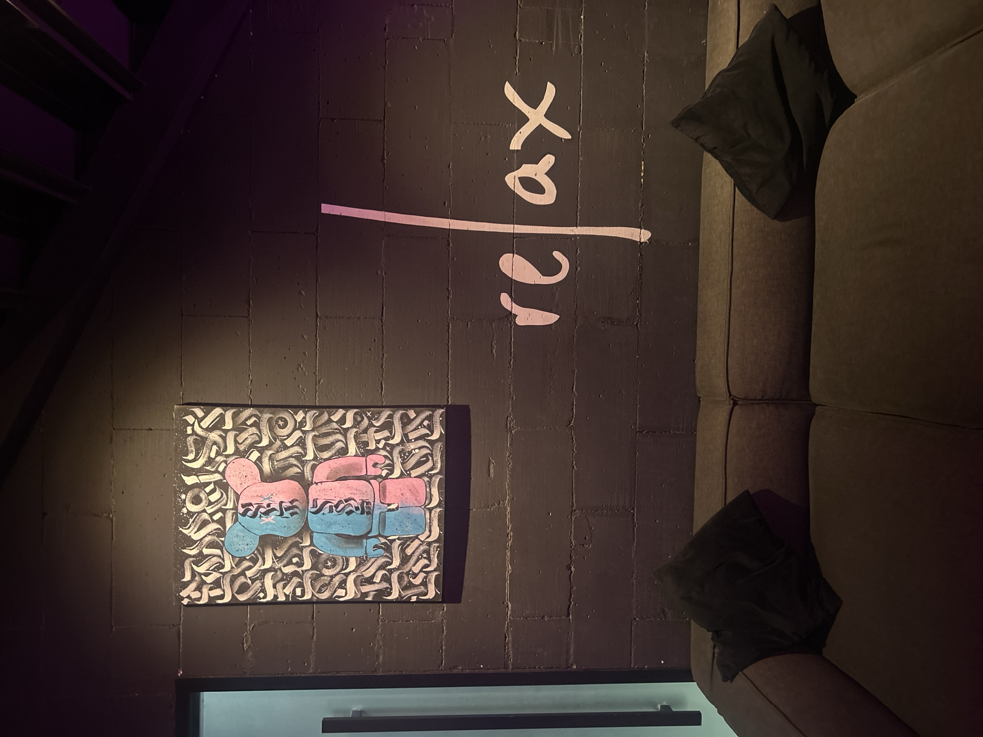 In North Kuta, Indonesia, a dark, textured brick wall serves as the backdrop for a modern, art-filled space. Mounted on the wall is a rectangular painting featuring a stylized bear figure, rendered in a split blue and pink color scheme against a chaotic, graffiti-like pattern in black and white. To the right of the painting, the word "relax" is stenciled onto the wall in a flowing, white script, accented by a vertical pink stripe that runs through the 'l'. Below the wall, a dark gray sectional sofa occupies the foreground, adorned with two black throw pillows. The lighting in the scene is dim, with a purple hue emanating from the upper right corner, casting subtle shadows and highlighting the textured surfaces of the wall and sofa. A sliver of a doorway with a vertical handle is visible on the far left, suggesting an interior space.